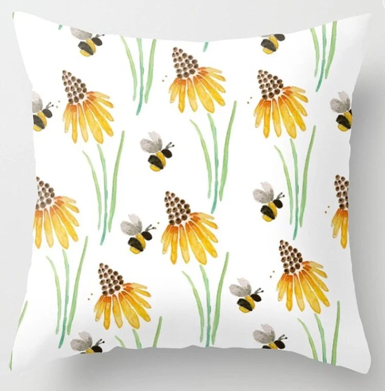 A decorative throw pillow with a white background, featuring a pattern of yellow-orange flowers, green stems, and bees with black and yellow striped bodies and gray wings.
