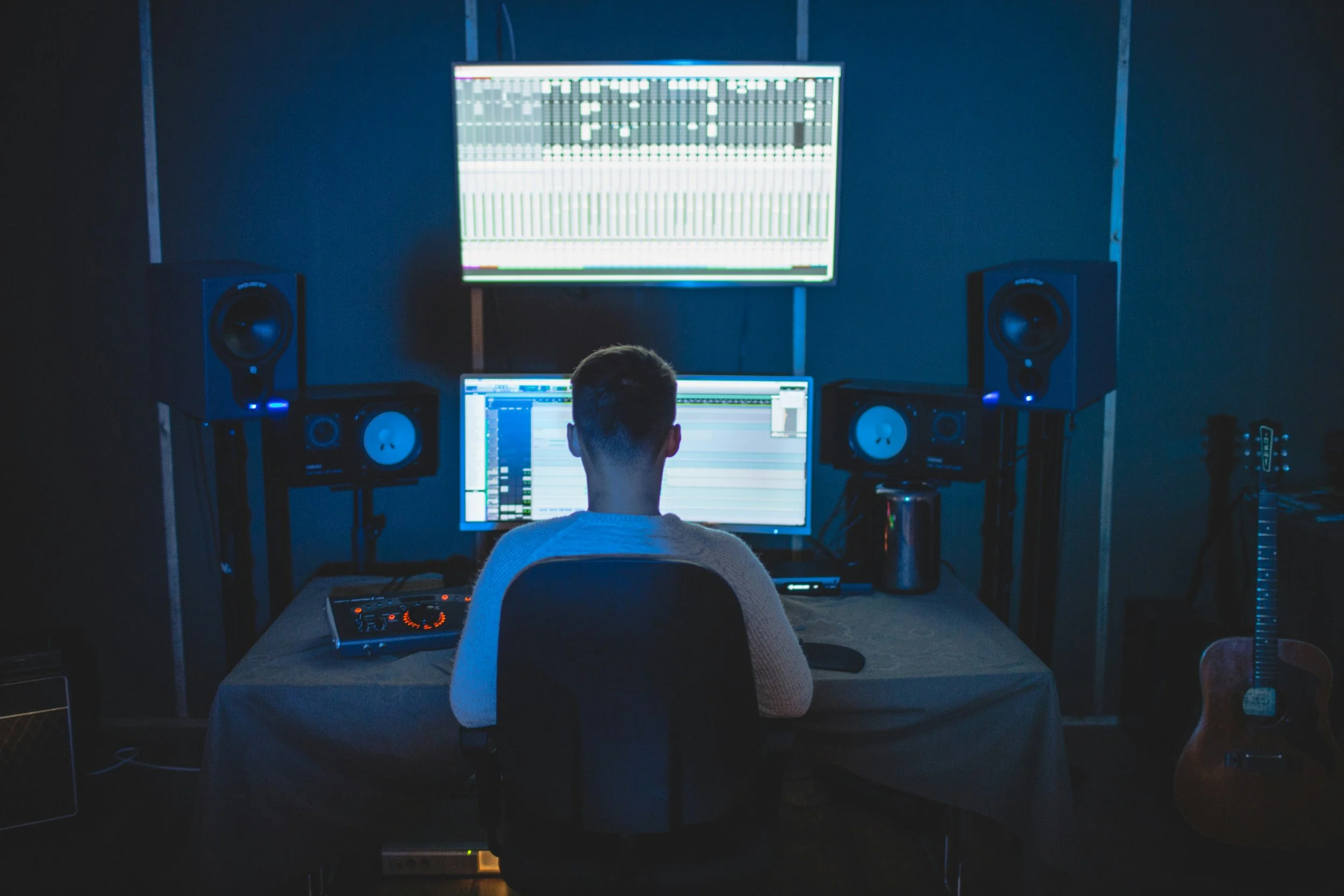 A person with short hair is sitting at a desk with music production equipment, including a computer, studio monitor speakers, and a MIDI keyboard. They are working on music software on the computer and looking at a large screen displaying digital audio workstations.