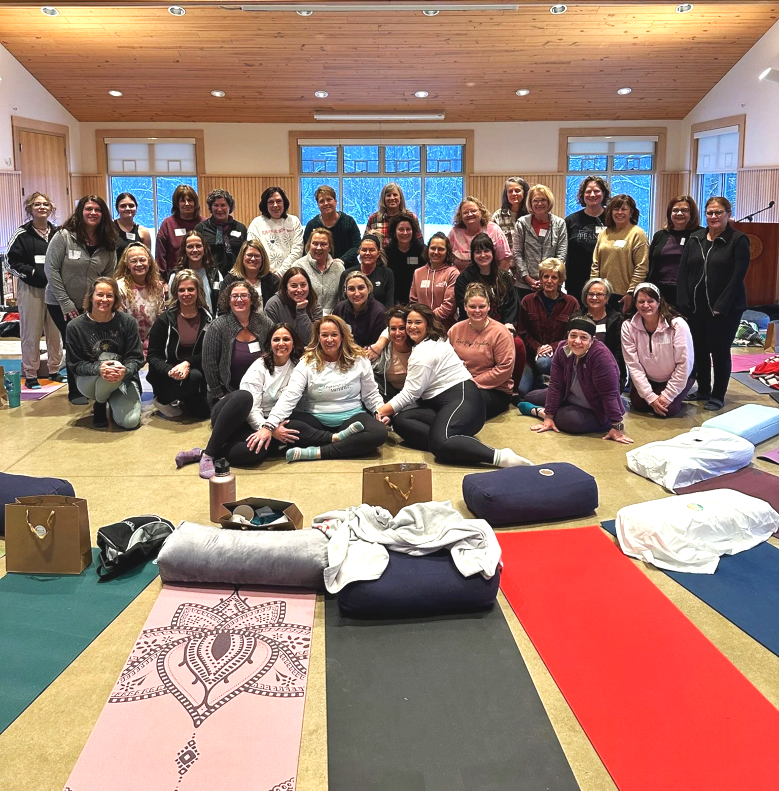 A group of women gathered together in a large indoor room with wooden paneling and windows showing a snowy landscape. They are posed for a group photo, some sitting on yoga mats and some standing behind. The room has yoga mats, blankets, and bags on the floor.