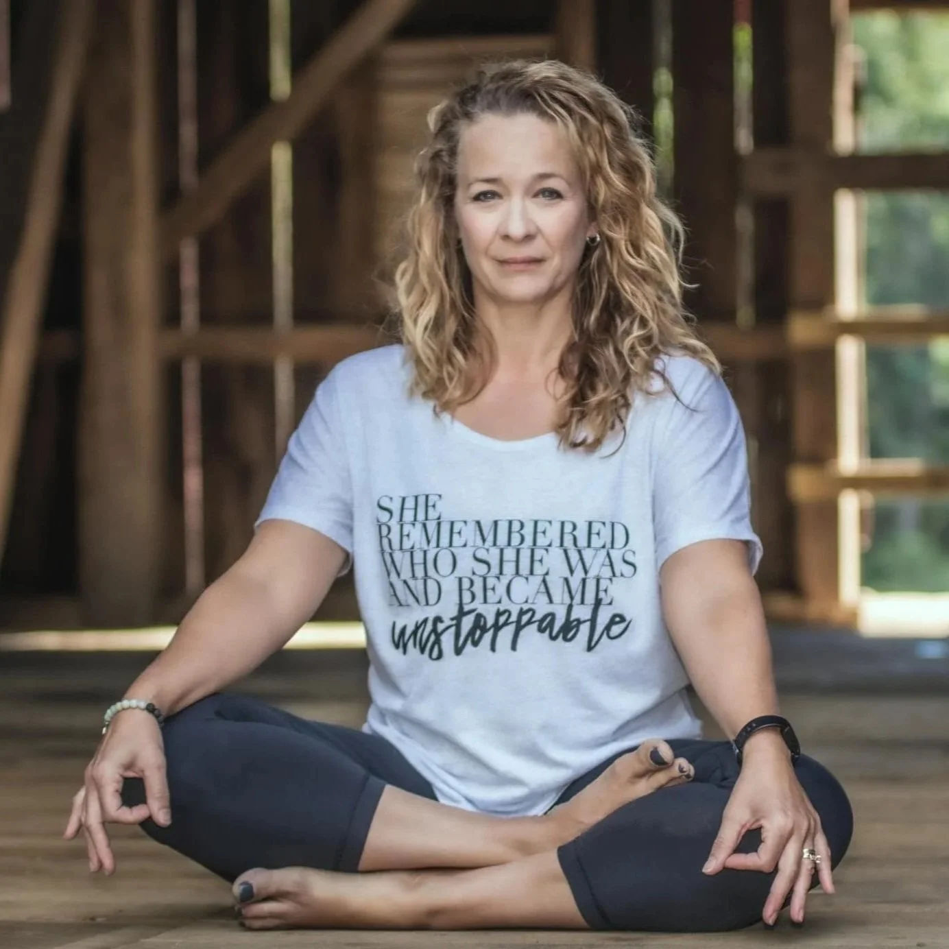 Woman sitting cross-legged on a wooden floor in a barn, practicing yoga, wearing a white T-shirt with text 'She remembered who she was and became unstoppable'.