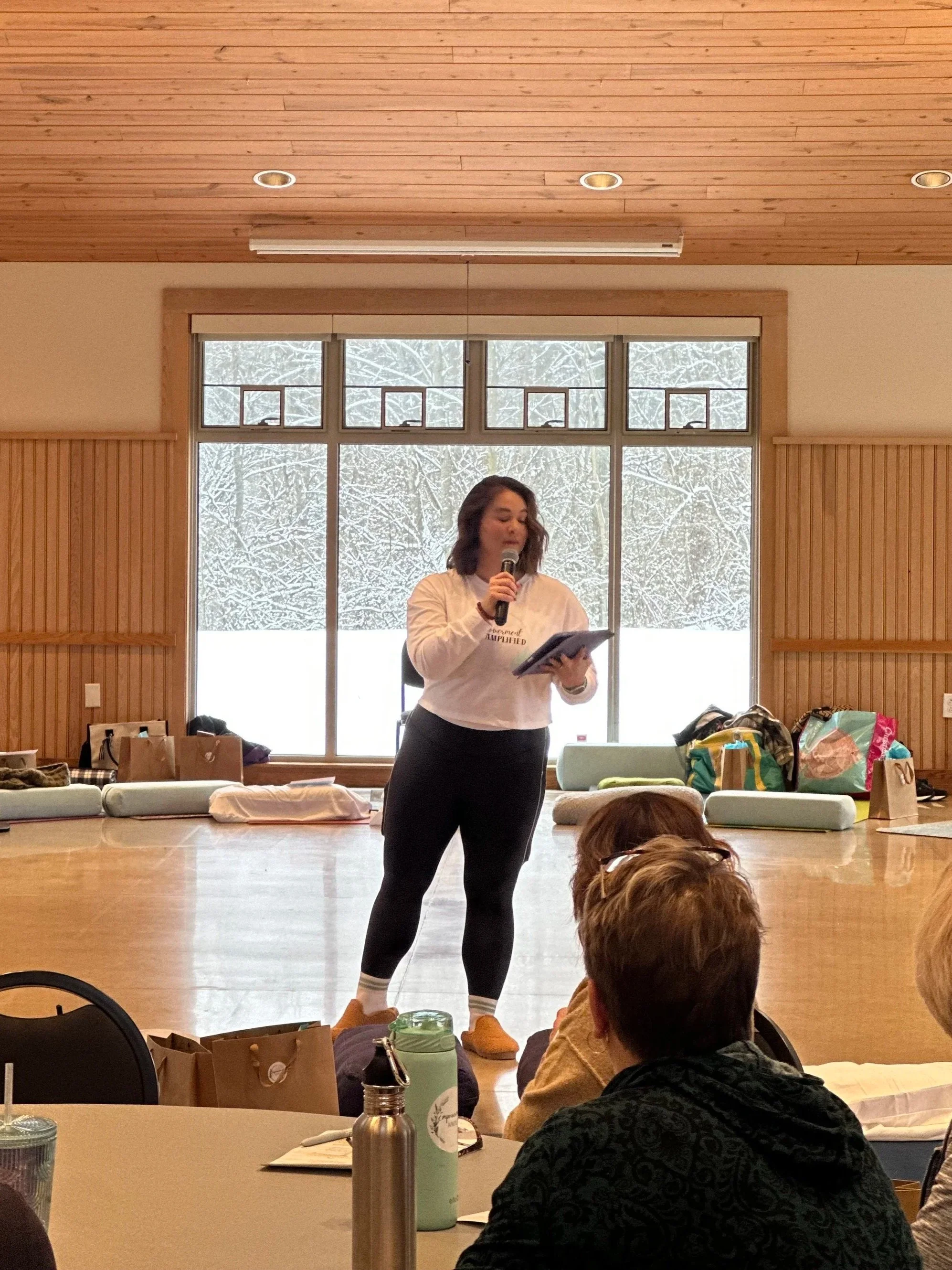 Woman standing and speaking into a microphone while holding a clipboard, in a room with large window overlooking snowy trees, surrounded by seated audience members, some with bags and drinks on table.