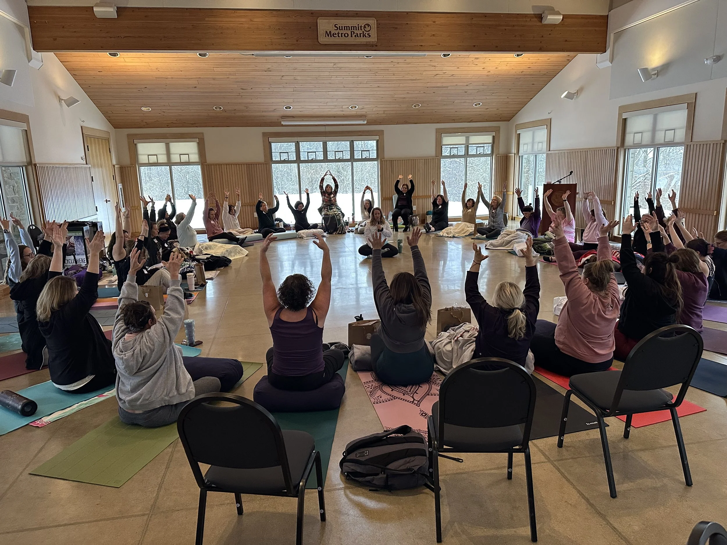 A large group of people participating in a yoga or meditation class inside a spacious room with natural light and wooden ceiling, with some sitting on mats with arms raised.