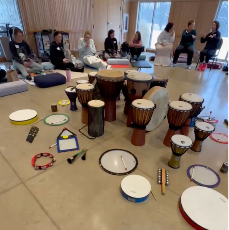 A group of people sitting on the floor in a room with large windows, with various drums and percussion instruments arranged in front of them.