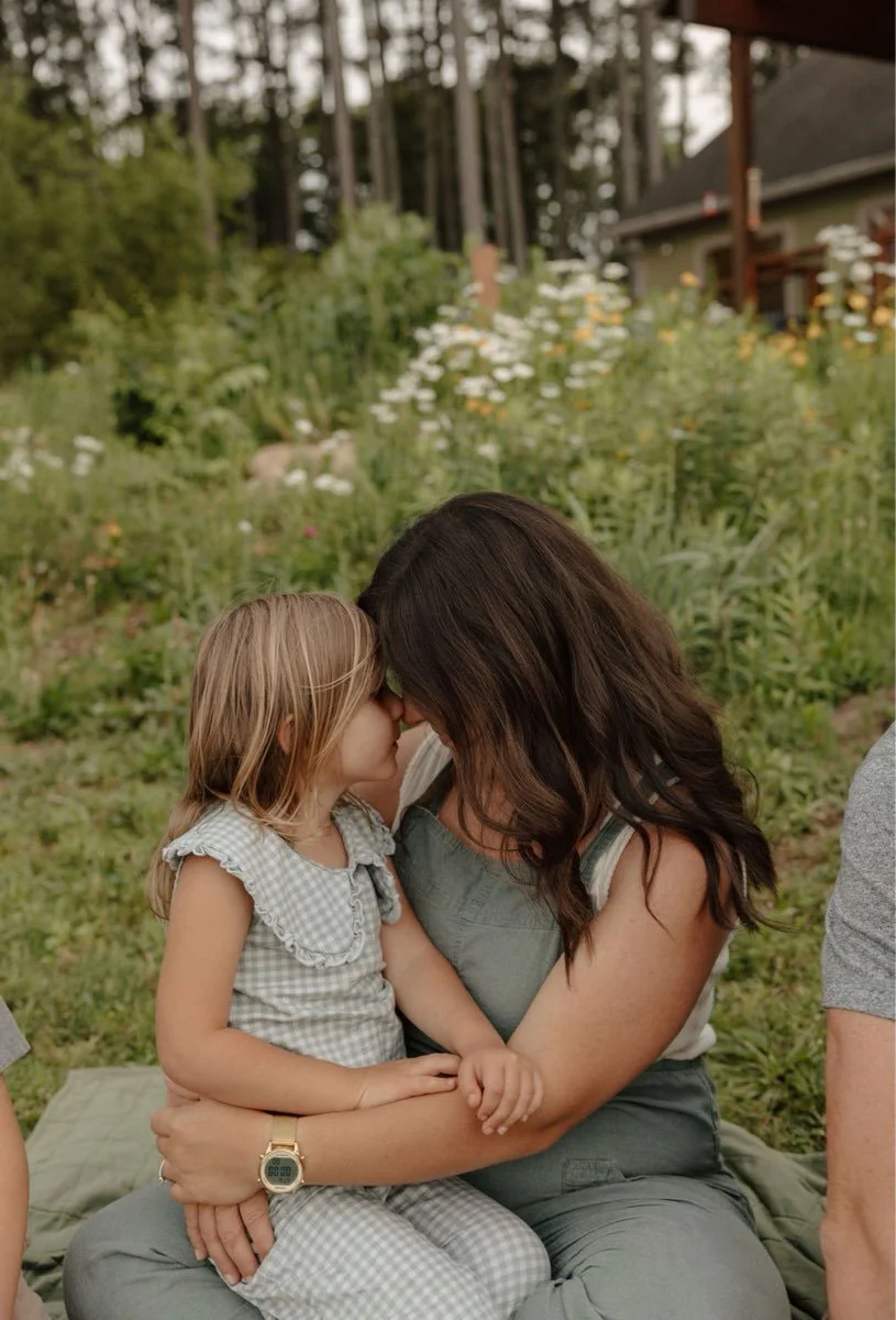 A woman and a young girl are sitting outdoors in a garden, touching foreheads lovingly with their eyes closed.