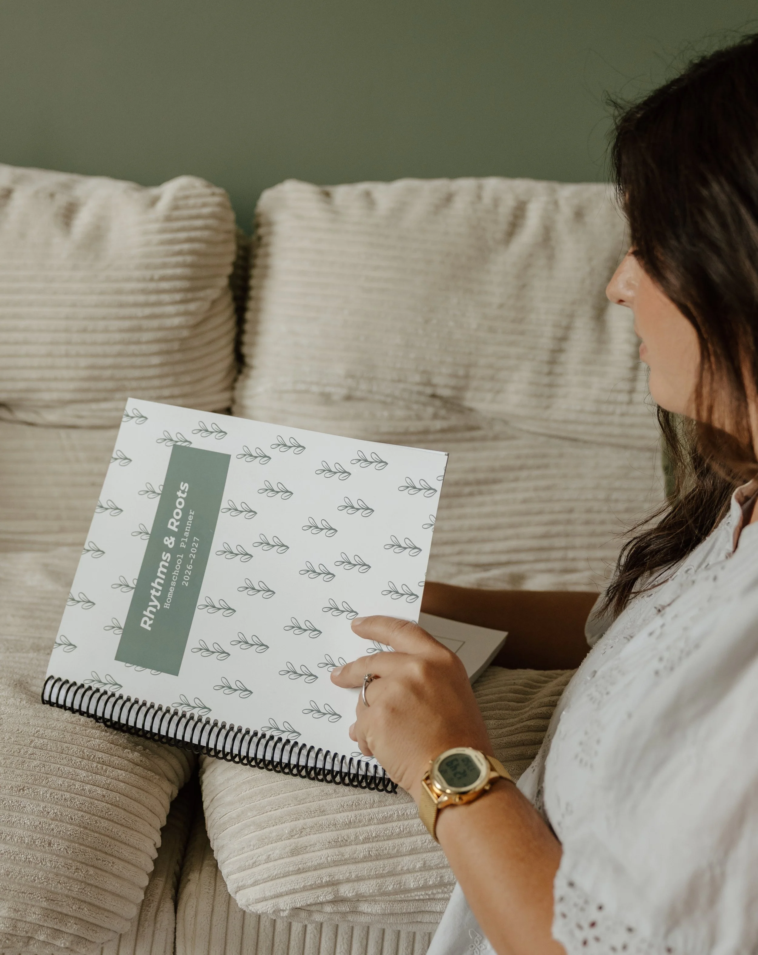A woman with dark brown hair, wearing a white lace blouse, gold watch, and rings, sitting on a beige corduroy couch, holding and reading a spiral-bound planner titled 'Rhythms & Roots' for the 2021-2022 academic year, with pillows behind her.