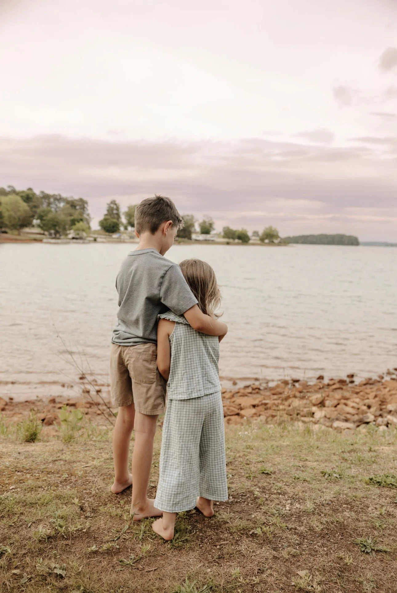 A boy and a girl standing by a lakeshore, embracing each other, at sunset with clouds and trees in the background.