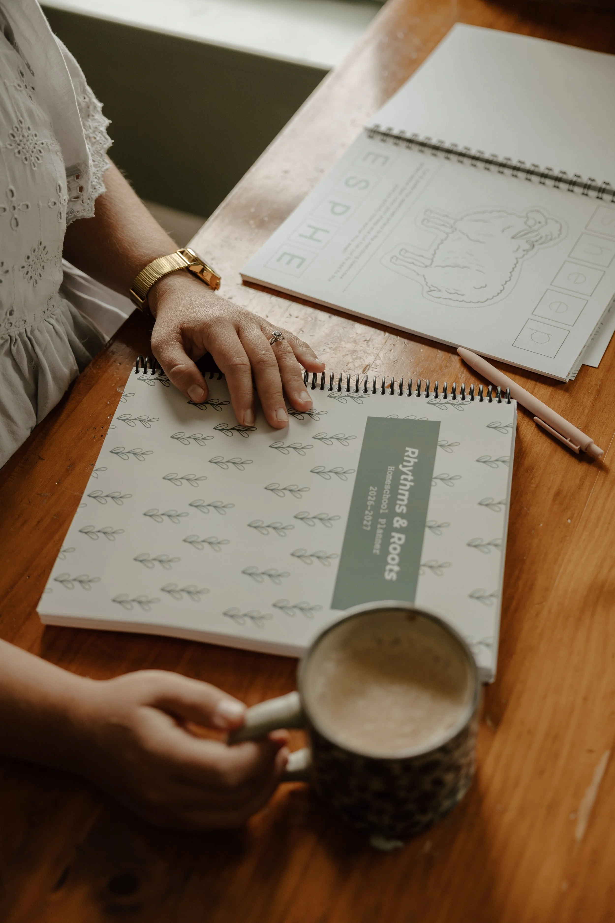 Person sitting at a wooden table with a workbook, a spiral-bound booklet, a pen, and a coffee mug.