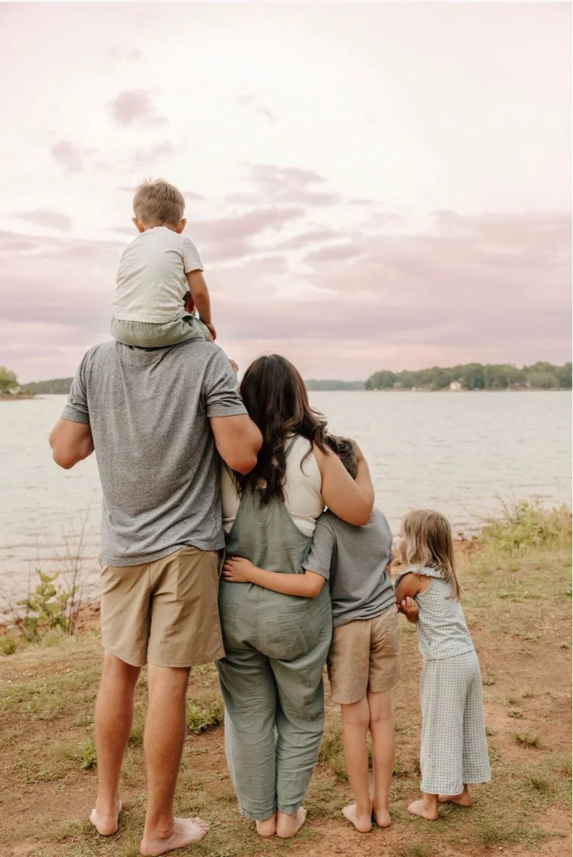 A family of five near a lake, with a man carrying a young boy on his shoulders while a woman and two children stand close by, all facing the water during sunset.