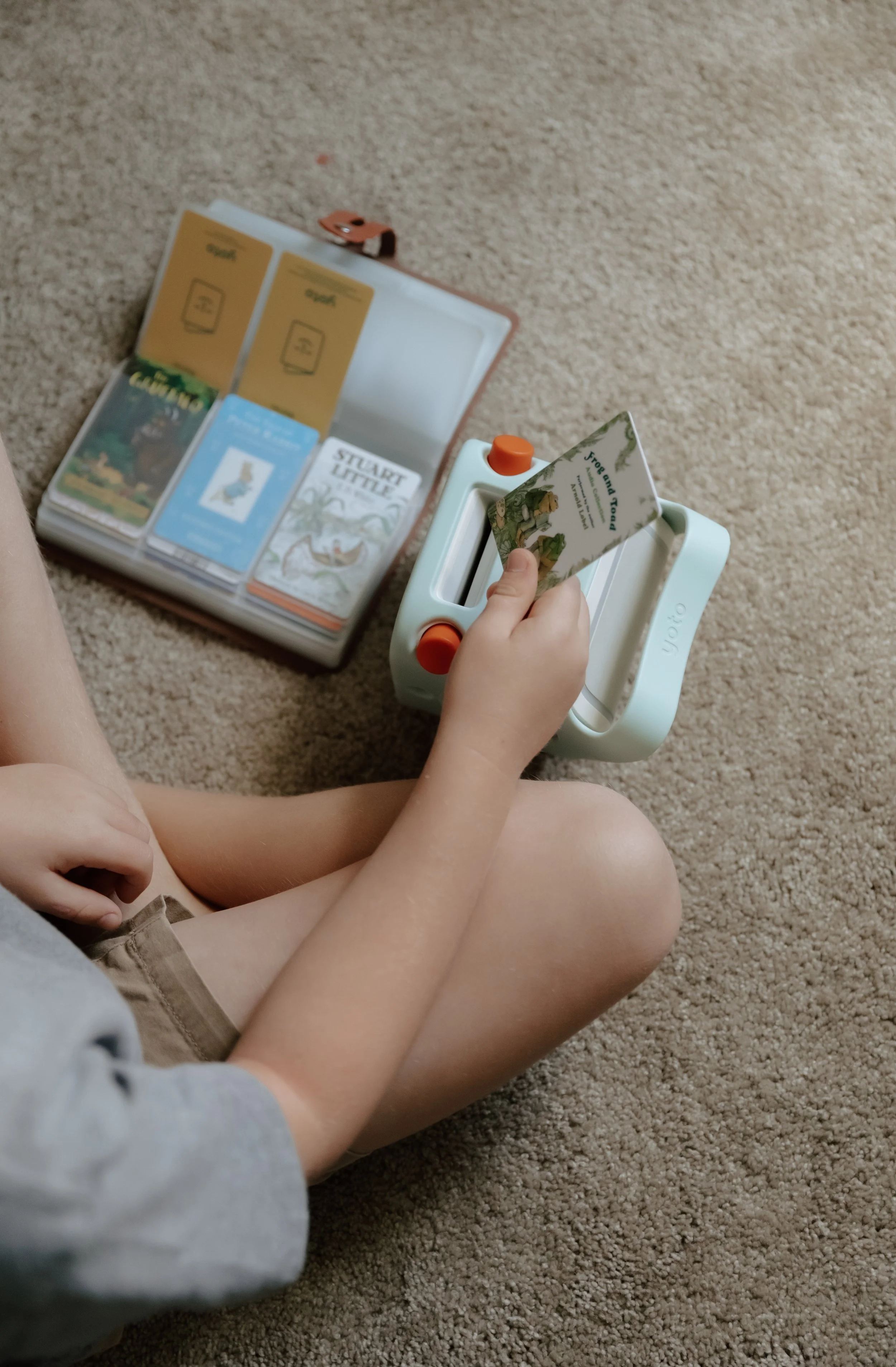 Child sitting on beige carpet, holding a card from a board game, with other game pieces and cards on the floor nearby.