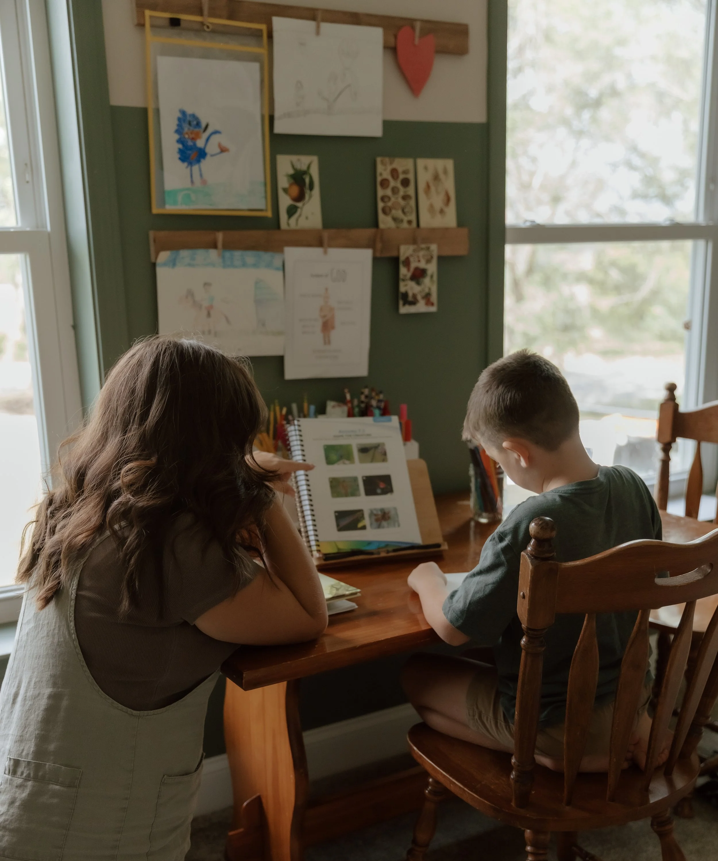 Two children sitting at a wooden table with a woman standing nearby, looking at an open book or scrapbook with colorful images inside. The room has a window and artwork hanging on the wall.