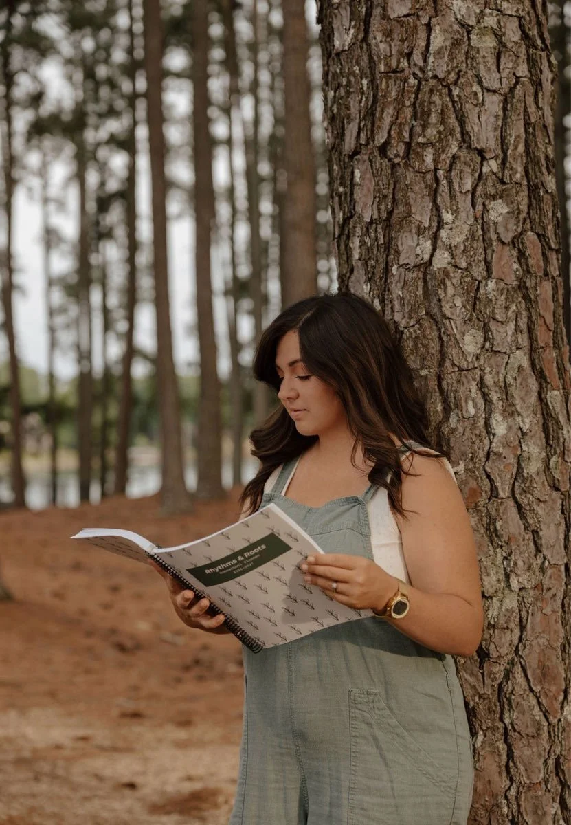 Young woman reading a book titled 'Rhythms & Rhymes' while leaning against a tree in a forested area.