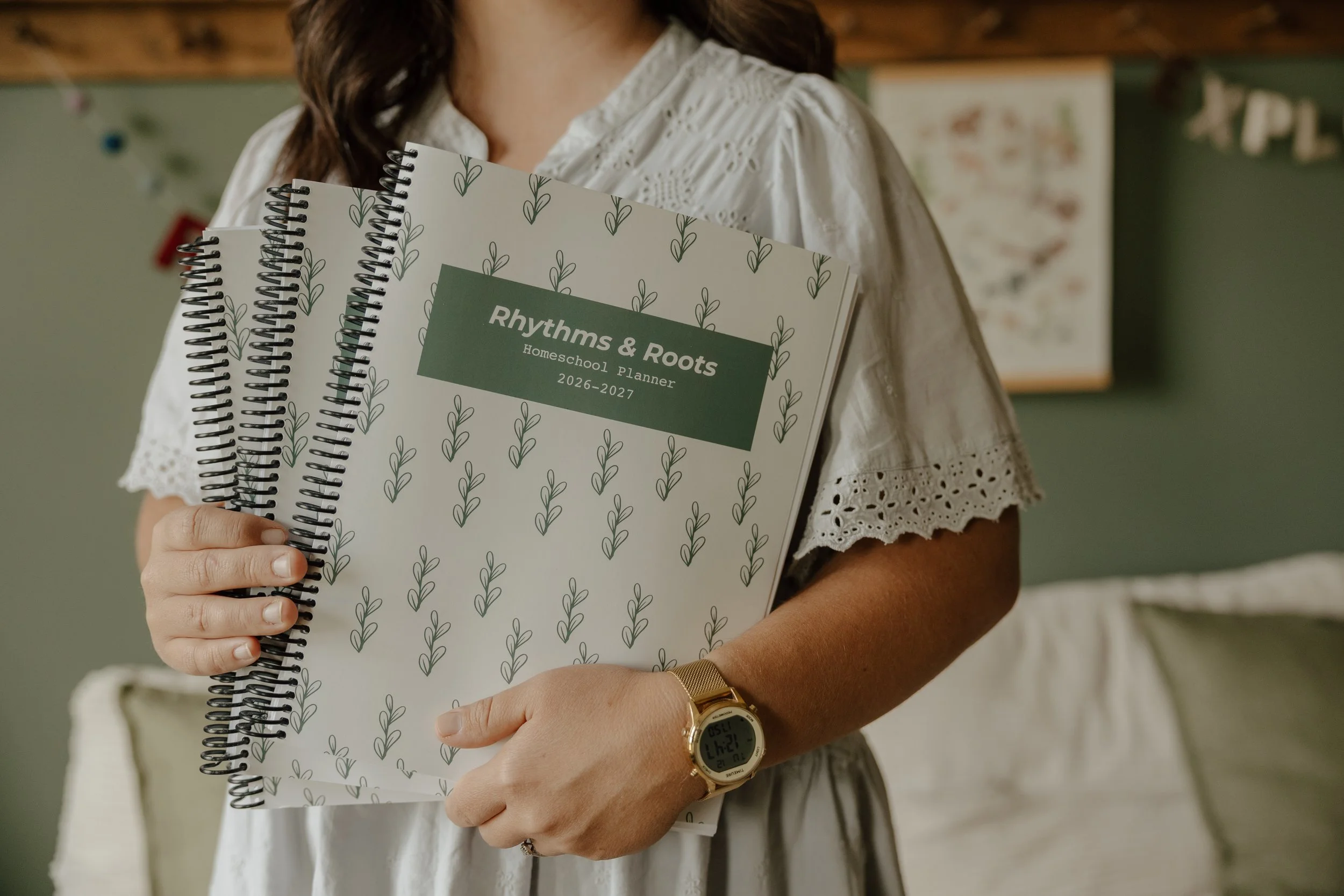 Person holding three planners titled 'Rhythms & Roots Homeschool Planner 2026-2027'. The person is wearing a white, embroidered blouse, a gold watch, and a gold bracelet. The background has a green wall with some children's artwork and decorations.