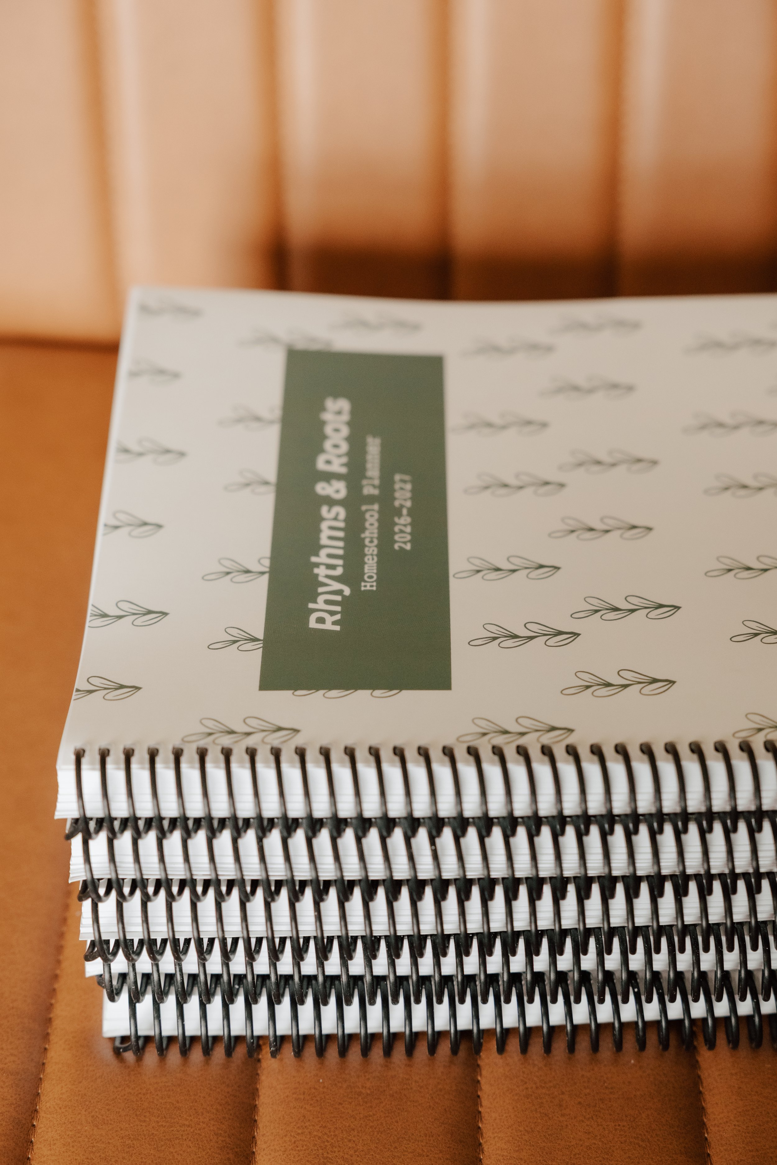 A spiral-bound notebook with a cover design of small leaves and a green rectangle in the center containing white text reading 'Rhythms & Roots', 'Homeschool Planner', and a date '2023-2024'. The notebook is placed on a wooden surface.