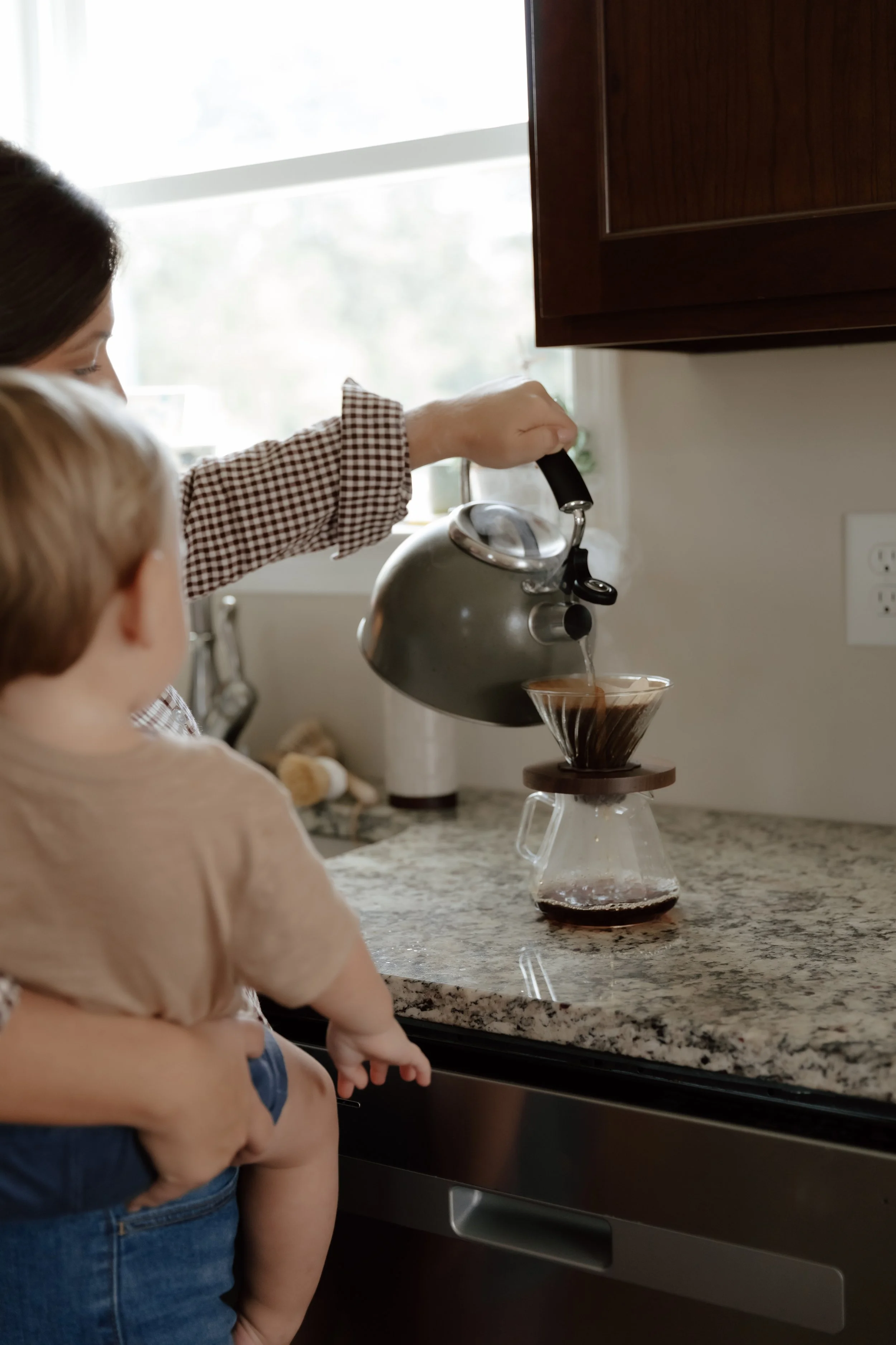 A woman is pouring hot coffee from a kettle into a glass coffee dripper, placed on a marble countertop with a young child beside her watching.