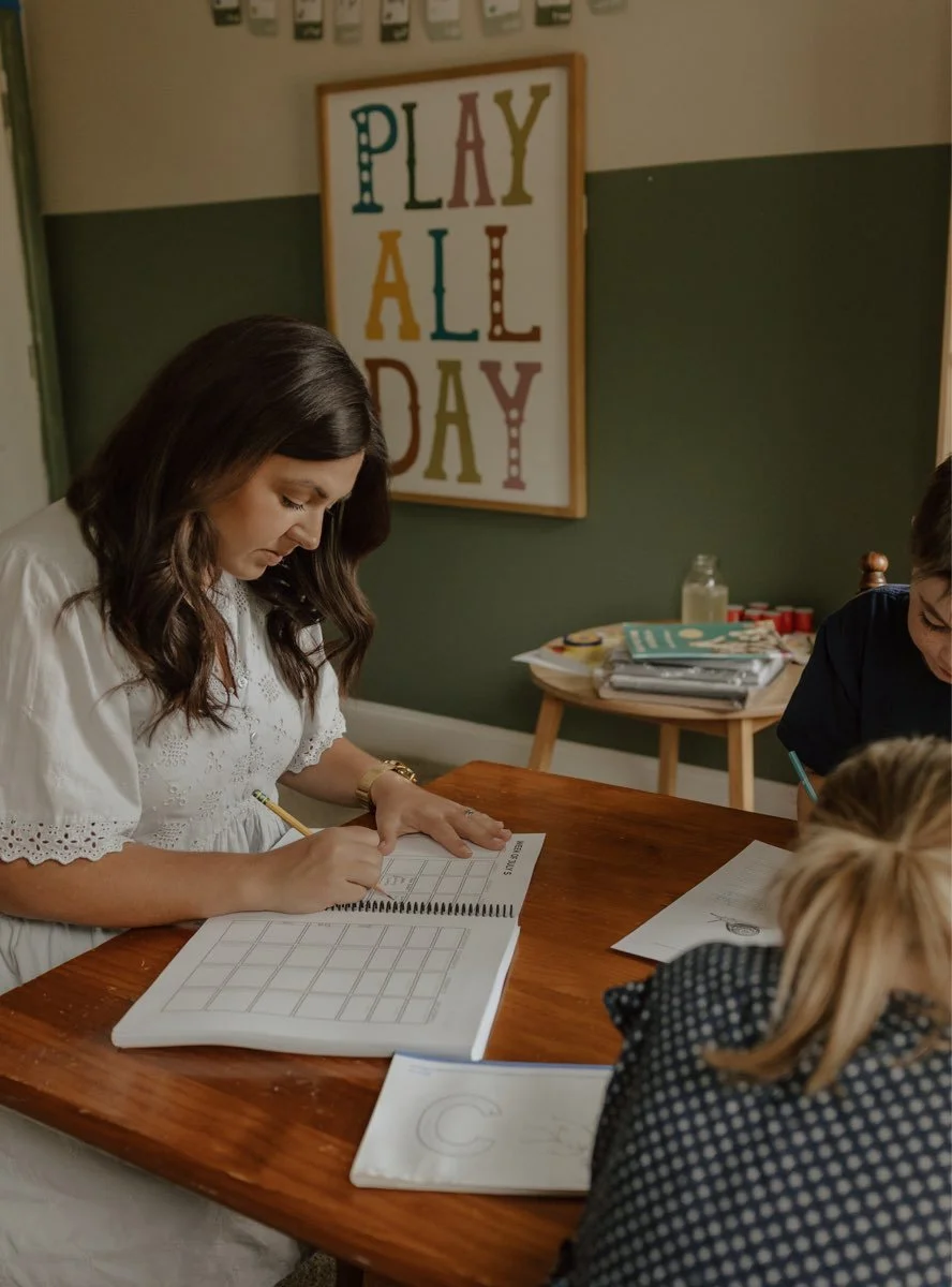Woman sitting at a wooden table writing in a spiral notebook with a group of children nearby, in a classroom with a colorful "PLAY ALL DAY" poster on the wall.