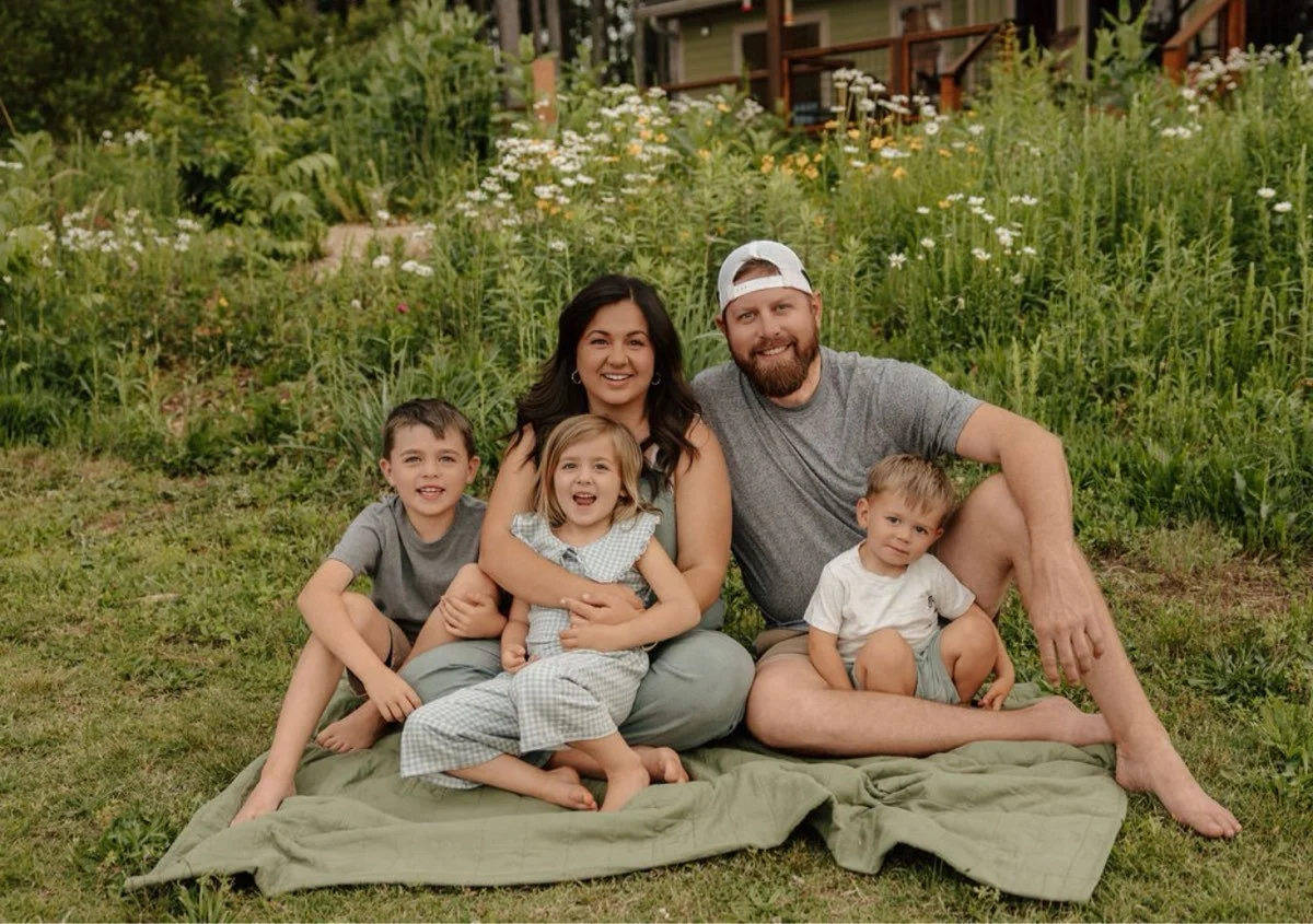 A family of five sitting on a blanket outdoors in a garden with flowers and greenery.