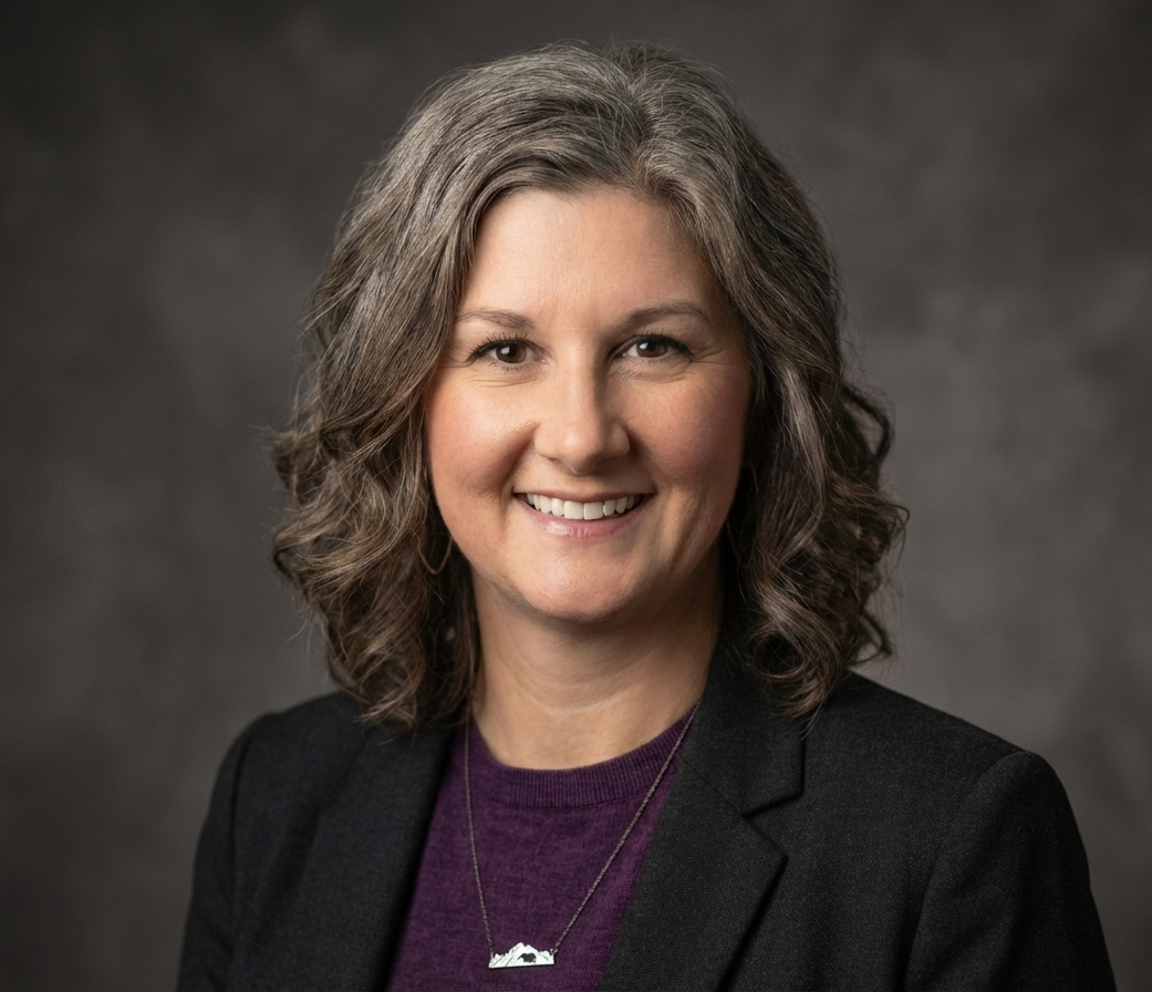 A professional woman with shoulder-length curly hair, smiling, wearing a dark blazer over a purple top and a silver necklace with a mountain and sunset pendant, against a gray background.