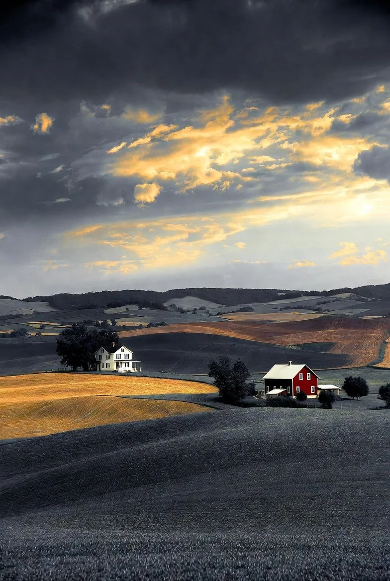 Scenic landscape with rolling hills, two houses (one white and one red), and a dramatic cloudy sky at sunset.