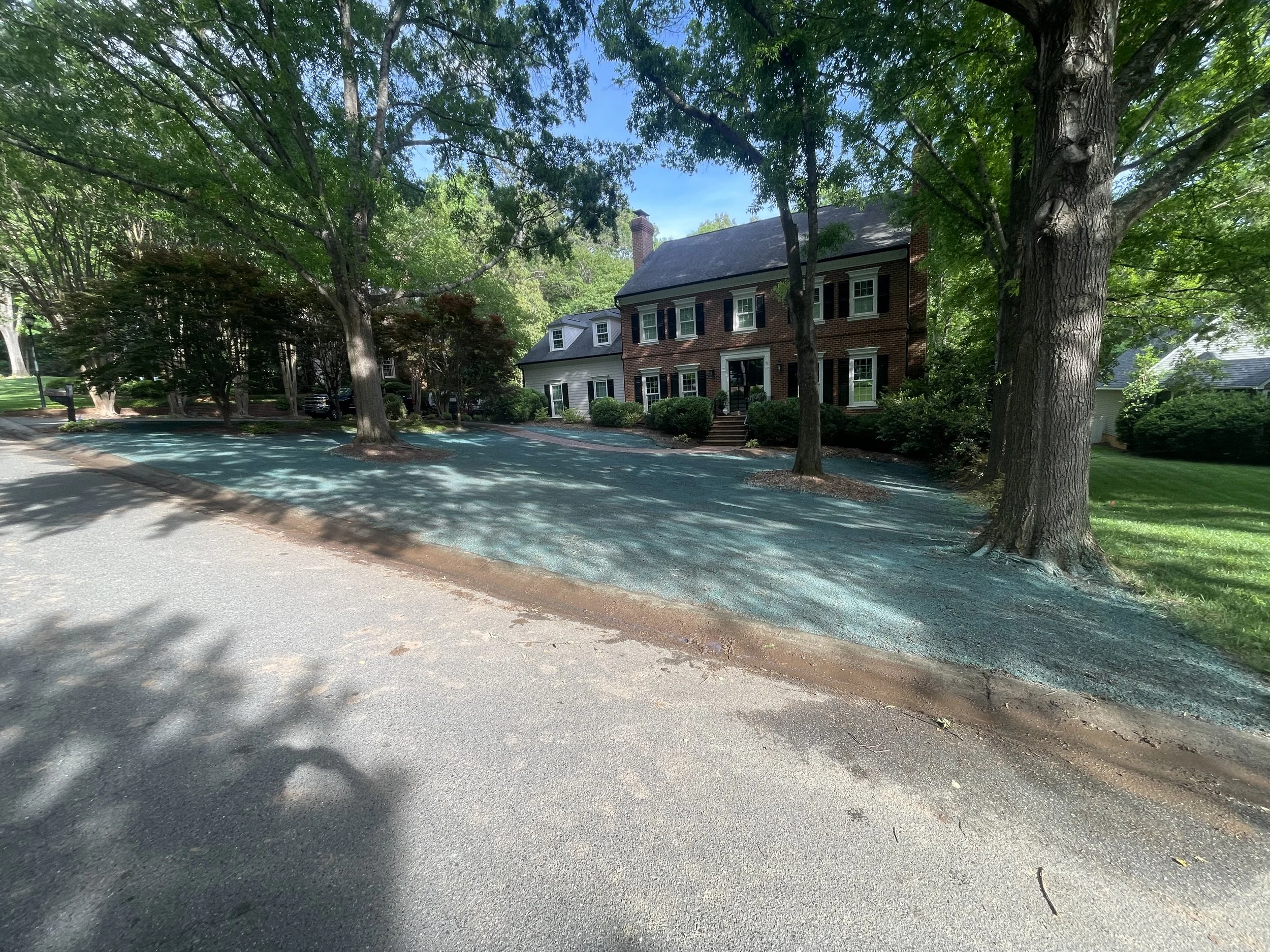 A residential neighborhood with a large brick house surrounded by trees and greenery. The street is shaded by trees and there are parked cars.