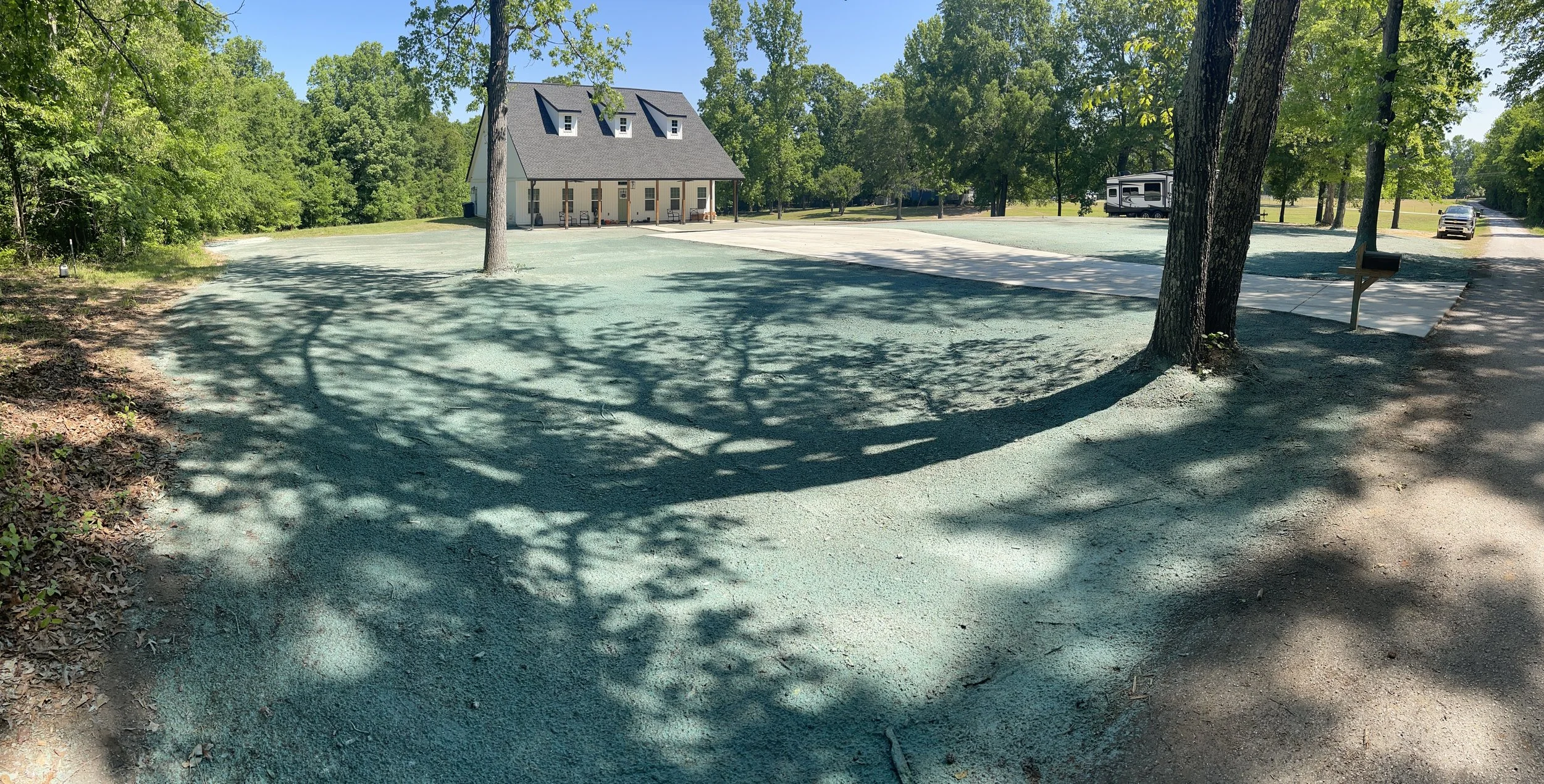 A gravel parking lot with a white building that has a porch and three small dormer windows on the roof. A black pickup truck and a white camper are parked in the background, surrounded by trees.