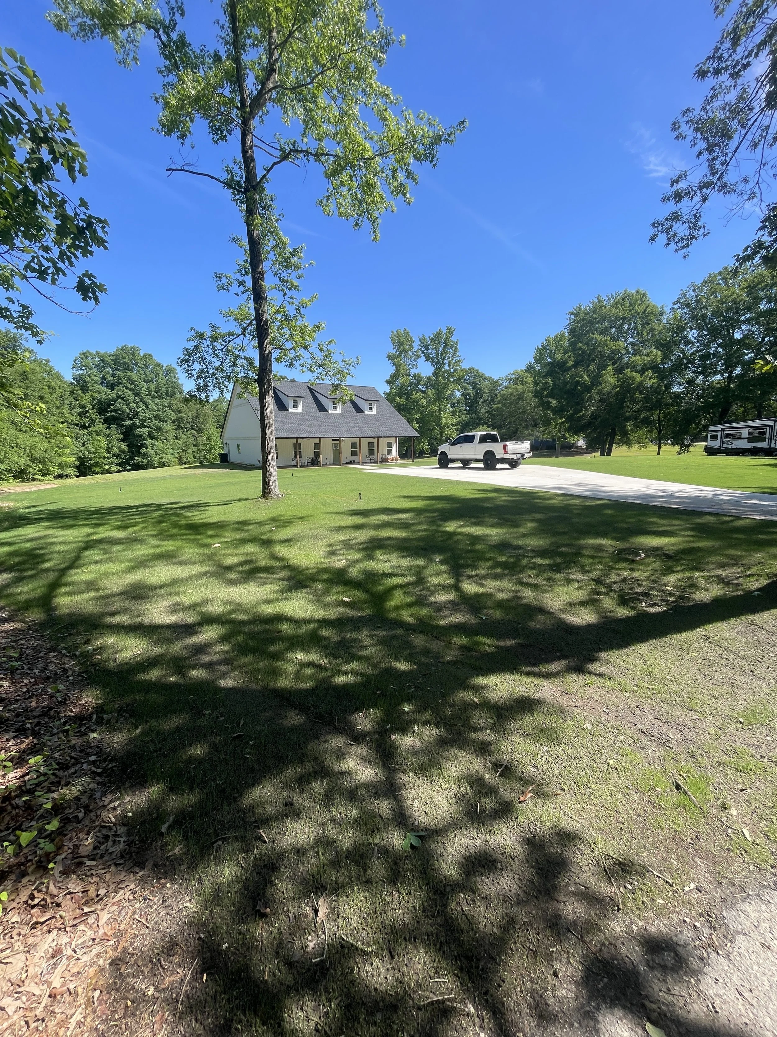 A white house with a gray roof, surrounded by green trees and grass, with a white truck and an RV parked on the driveway under a bright blue sky.