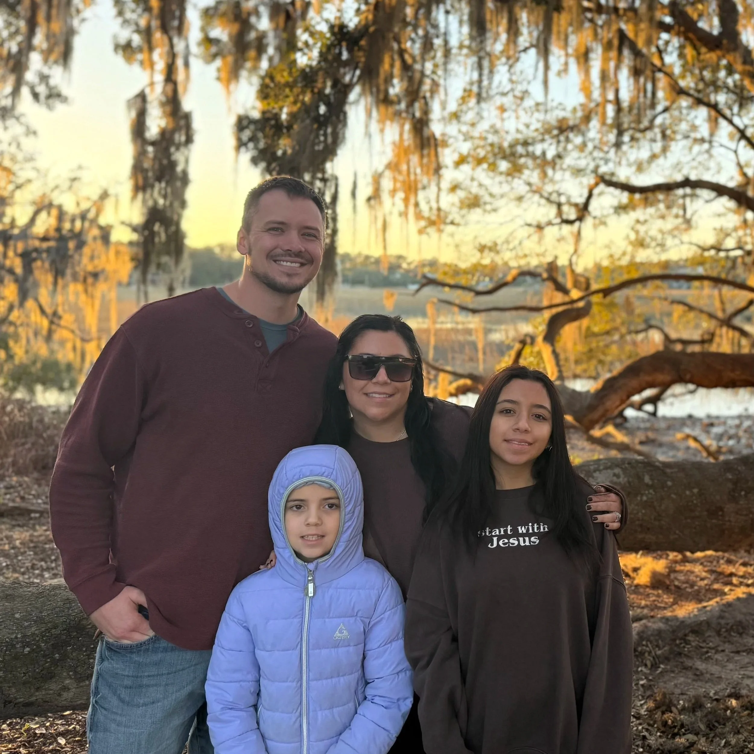 A group of four people outdoors during autumn, standing together in front of a tree with yellow and orange leaves. The group includes two women and two men, smiling at the camera. One woman is wearing sunglasses, and one person is wearing a light blue hoodie. The background shows a body of water and a sunset sky.