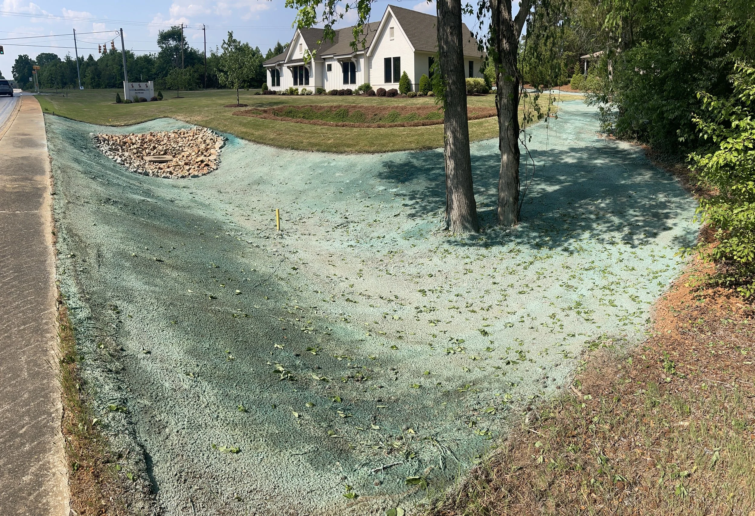 A construction zone with a green gravel pathway, a pile of rocks, and trees. There is a house in the background and an asphalt road on the left with a car and traffic light.