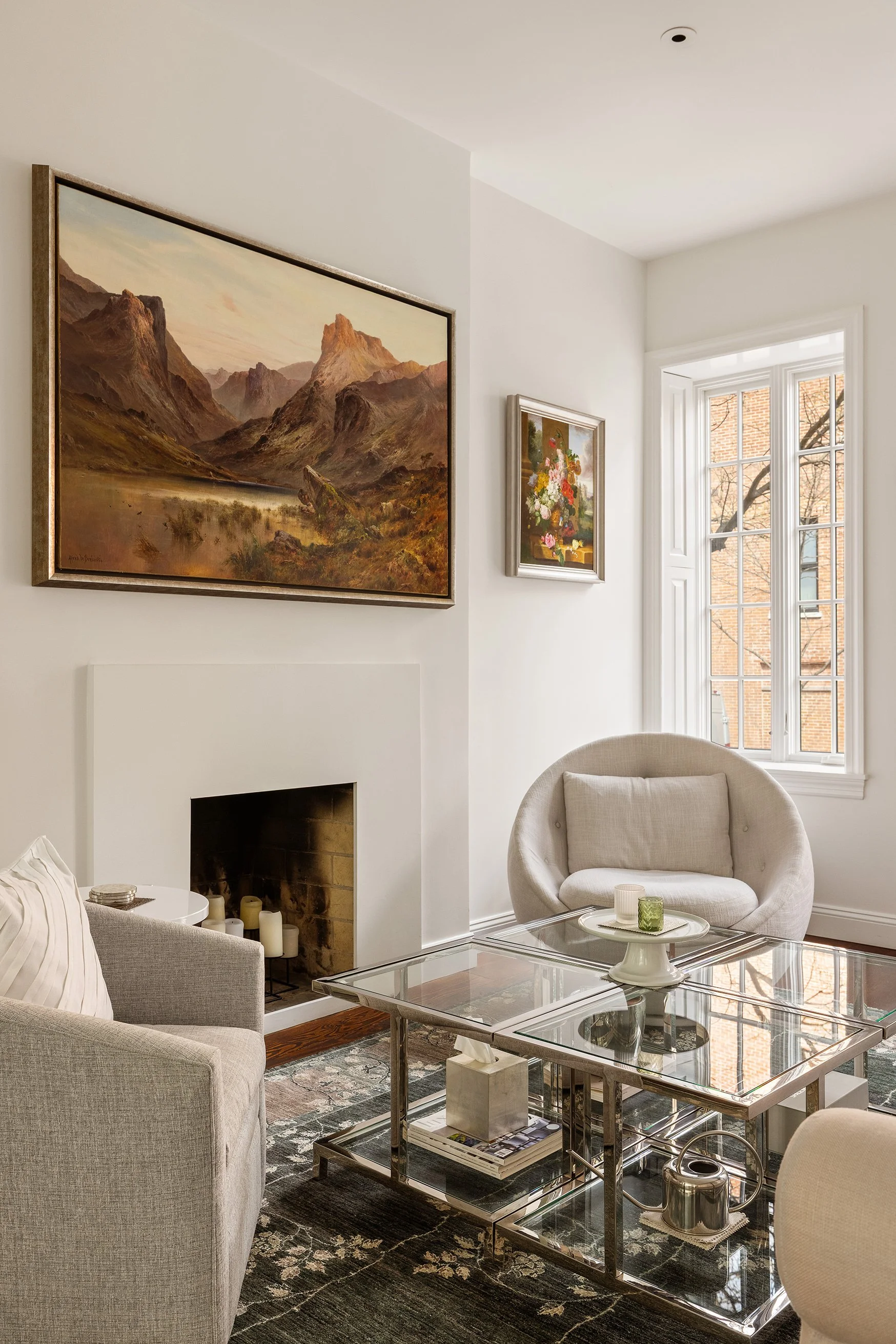 A cozy living room with a white fireplace, a glass coffee table, and a white round armchair near a large window. There are three paintings on the wall, including a landscape and a floral still life.