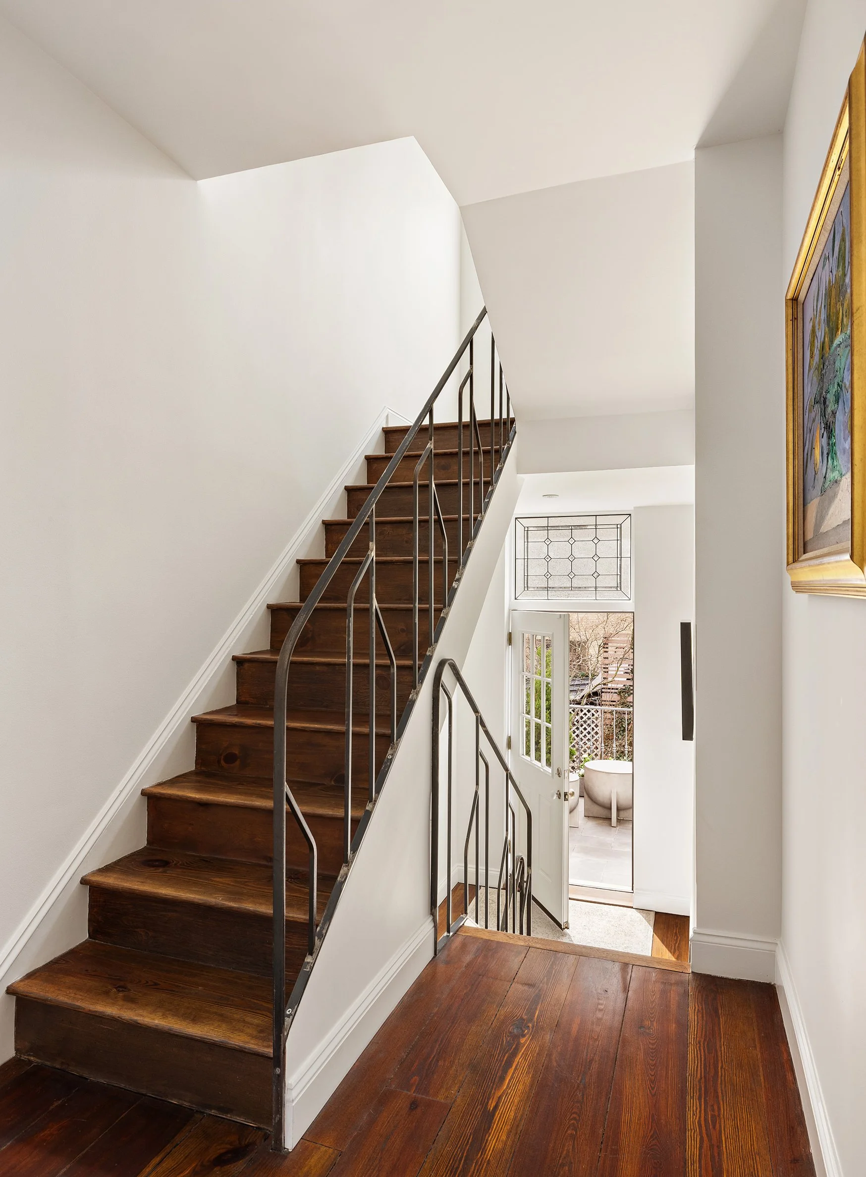 Interior view of a staircase with wooden steps and black metal railing, leading to an outdoor patio area with a door and windows. The space has white walls and hardwood flooring.