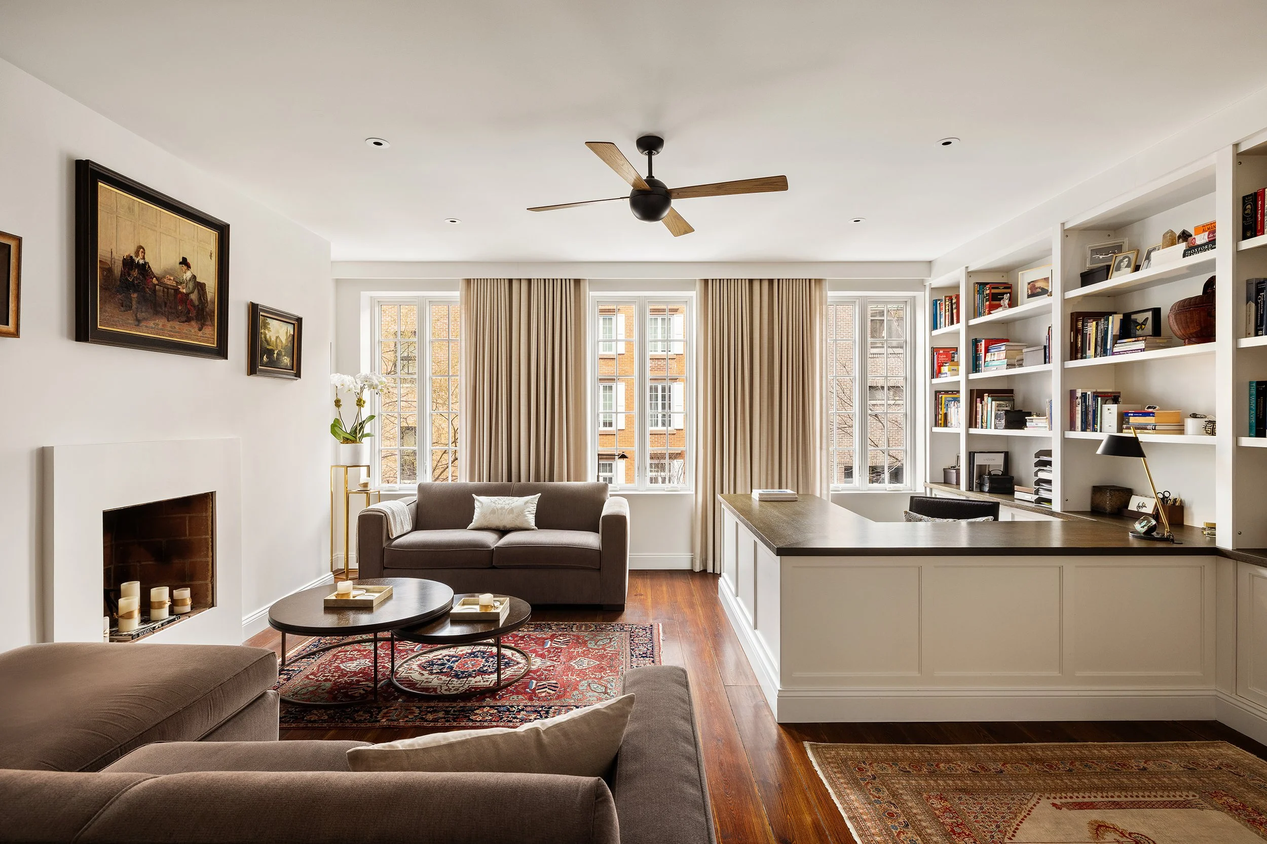 Bright living room with large windows, beige curtains, a bookshelf, a gray sofa with a pillow, a fireplace with candles, a ceiling fan, and a patterned rug.