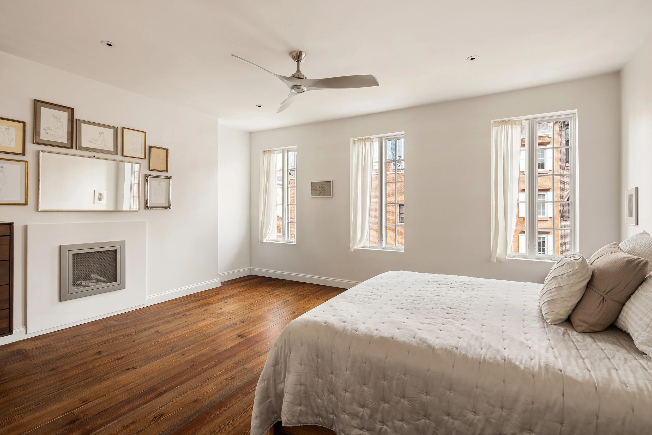 Bright bedroom with white walls, wood floors, a white bed with pillows, windows with white curtains, a ceiling fan, and framed artwork on the wall.