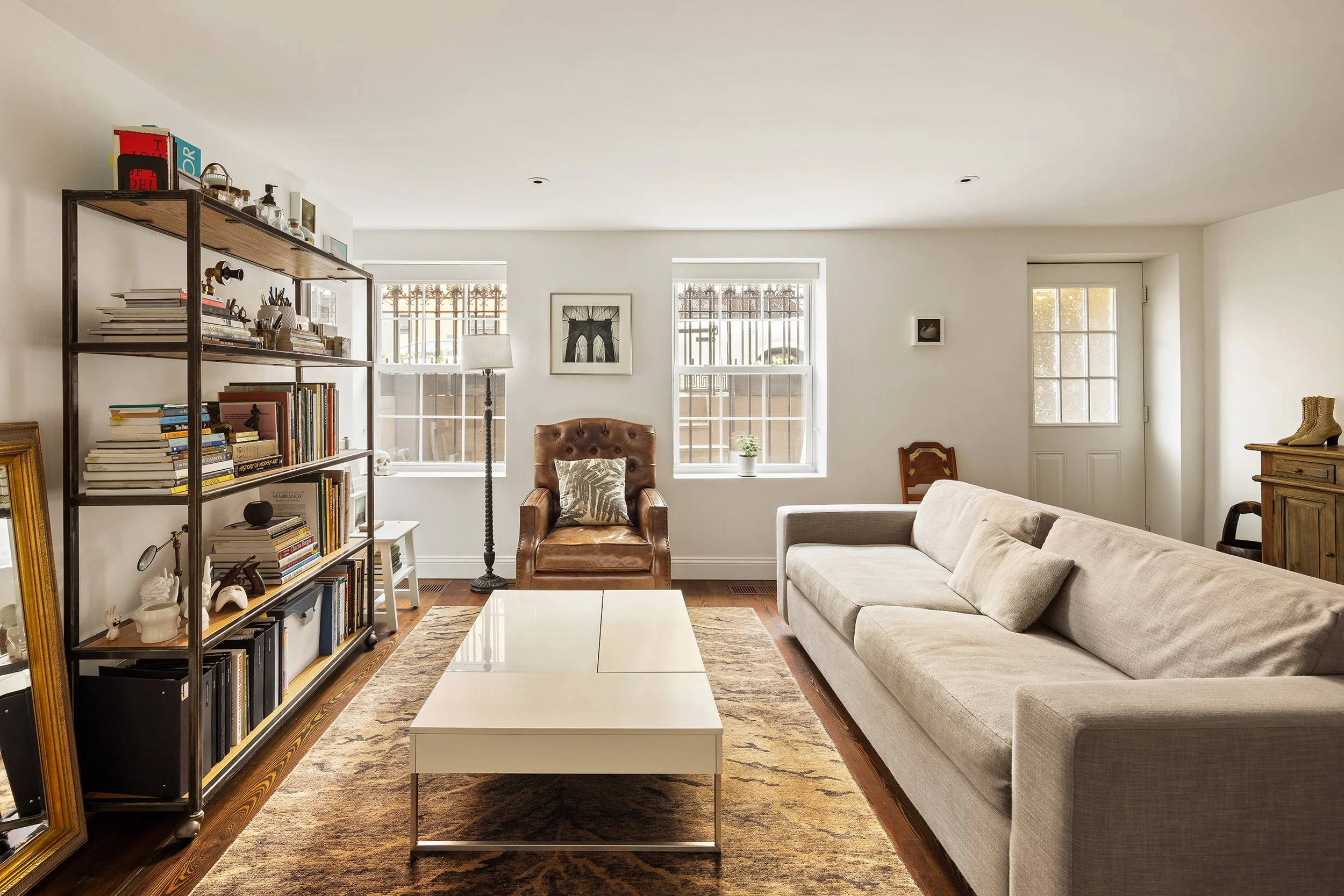 Living room with a beige sofa, a brown leather armchair, a white coffee table, a metal bookshelf filled with books and decor, a large rug, two windows with blinds, and a door by the window.