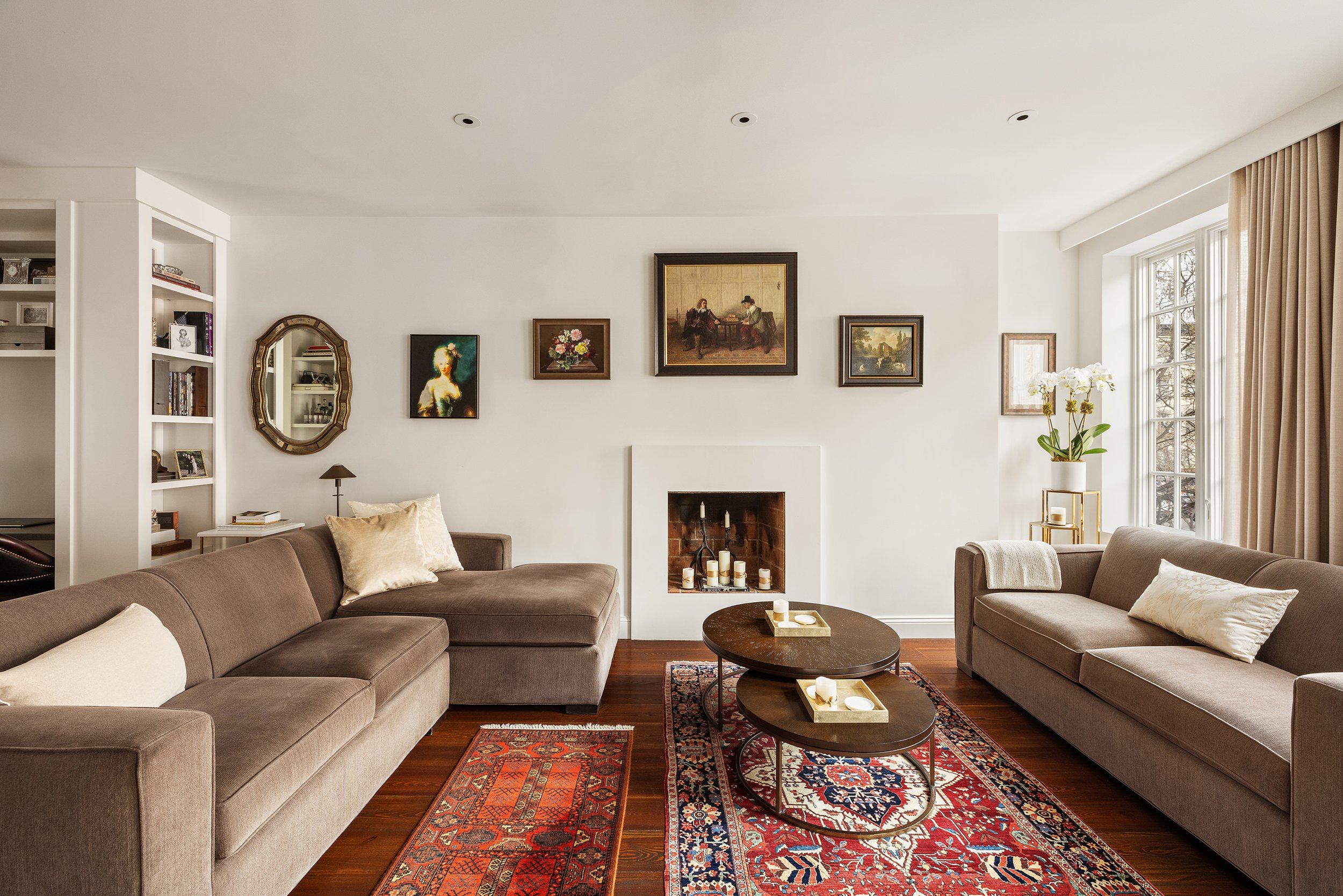 Living room with two beige sofas, a round coffee table with trays, a patterned rug, a fireplace, wall art, a mirror, a window with curtains, and a potted plant.