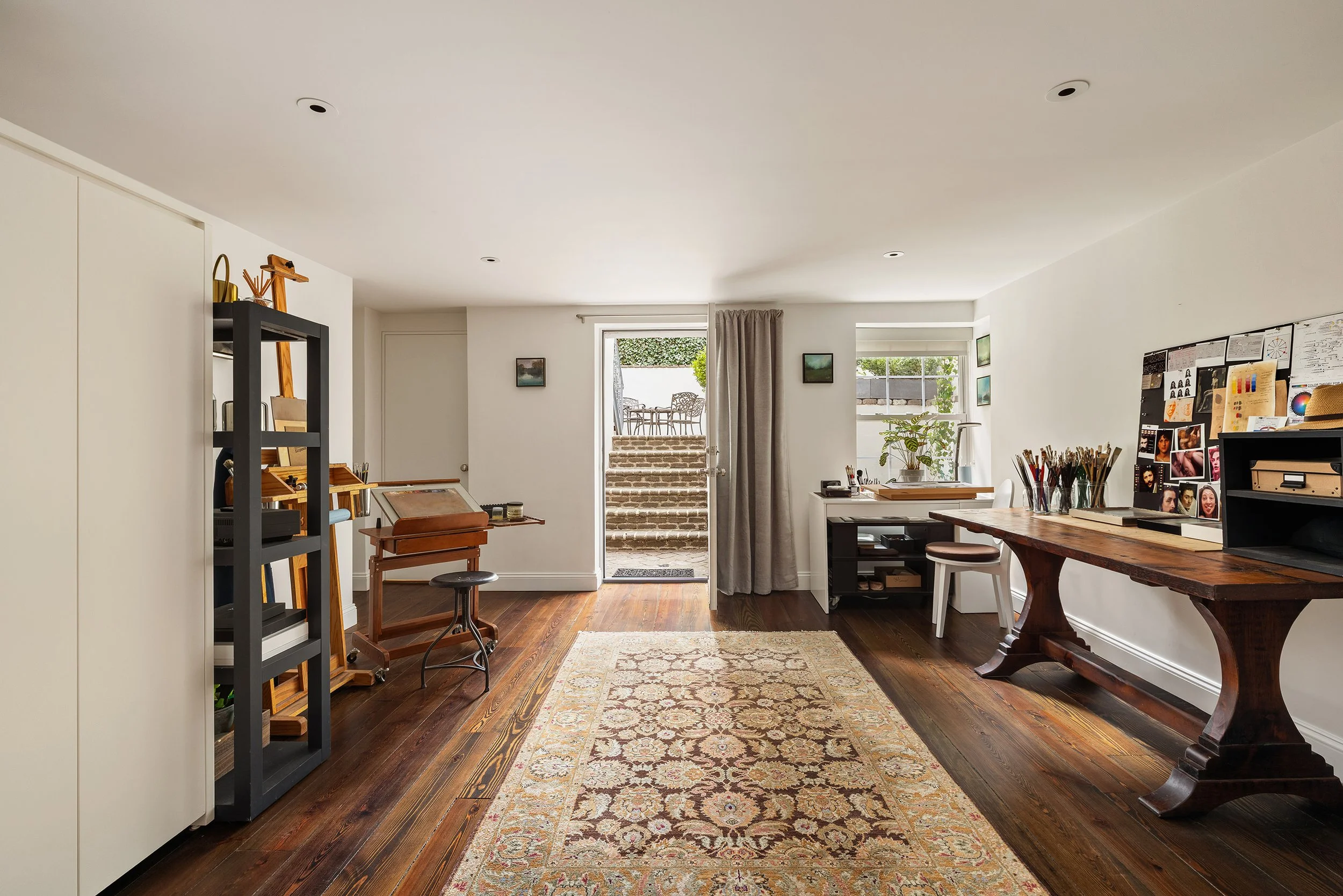 Living room with wooden floor, bookshelf, art supplies, desk, patterned rug, and large windows with curtains leading to an outdoor patio.