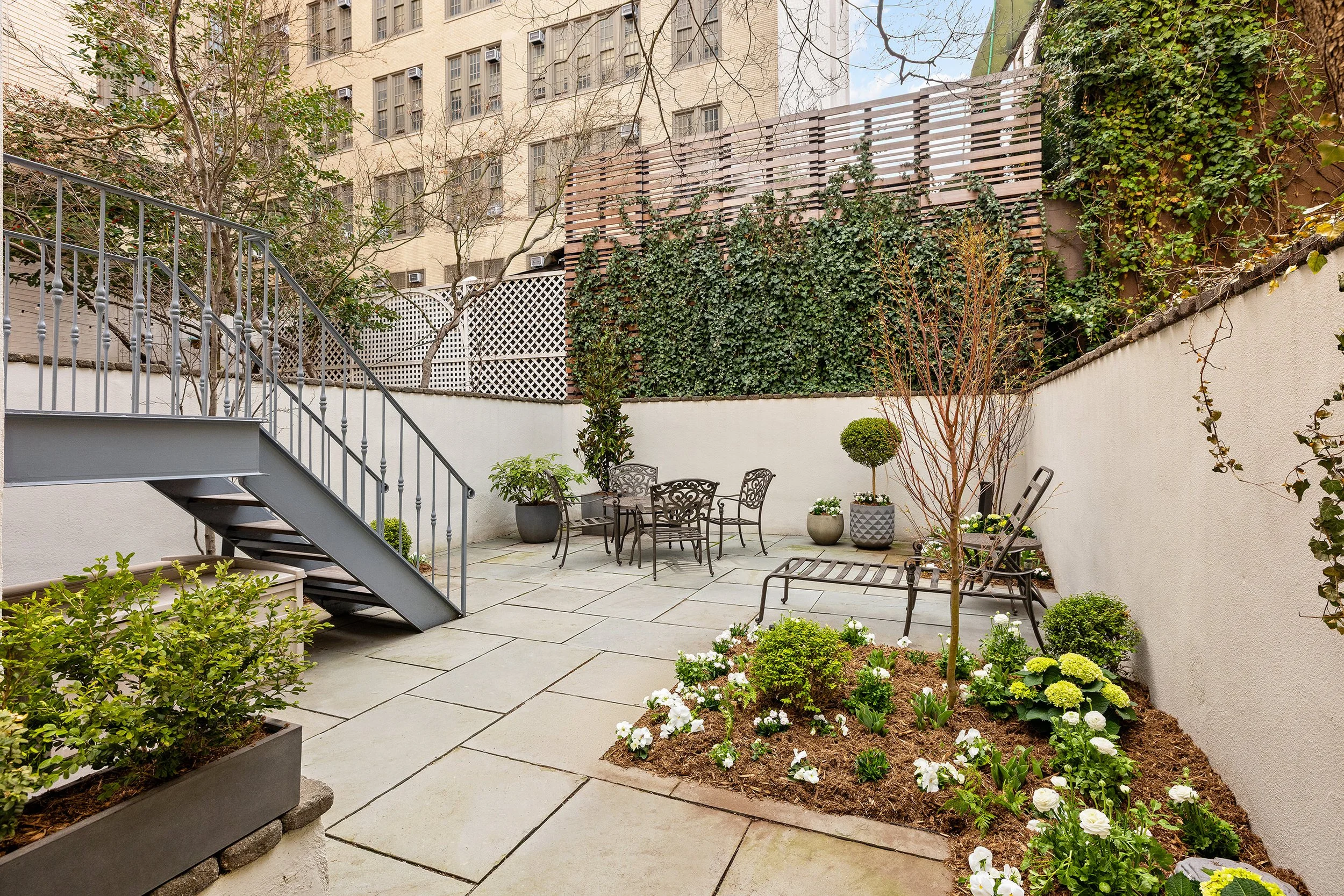 An outdoor patio with patio furniture, potted plants, and a small garden bed with blooming white flowers, surrounded by a high wall with climbing ivy and a wooden fence.