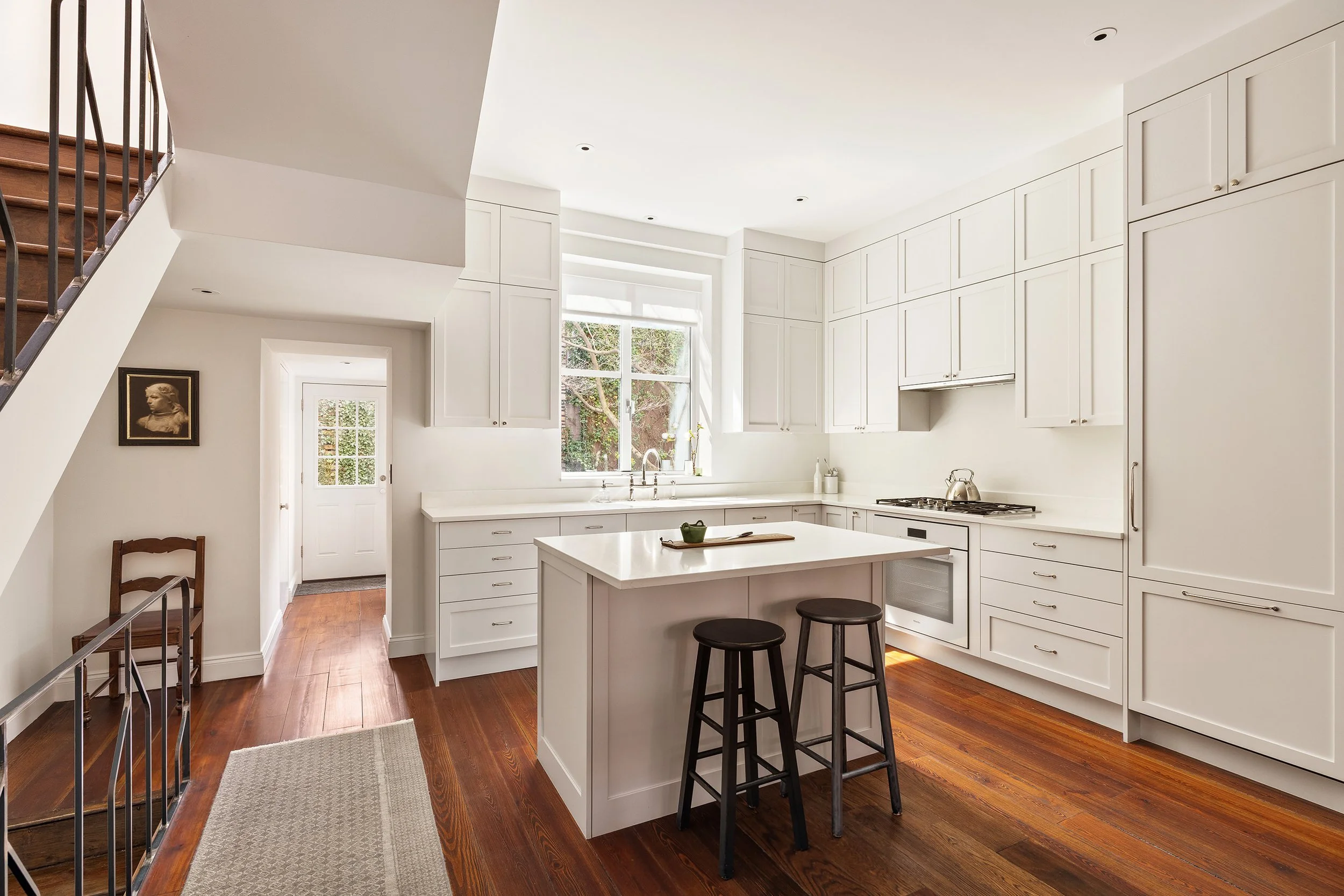 Bright white kitchen with island, two black stools, wooden floors, large window, white cabinets, and stainless steel appliances.