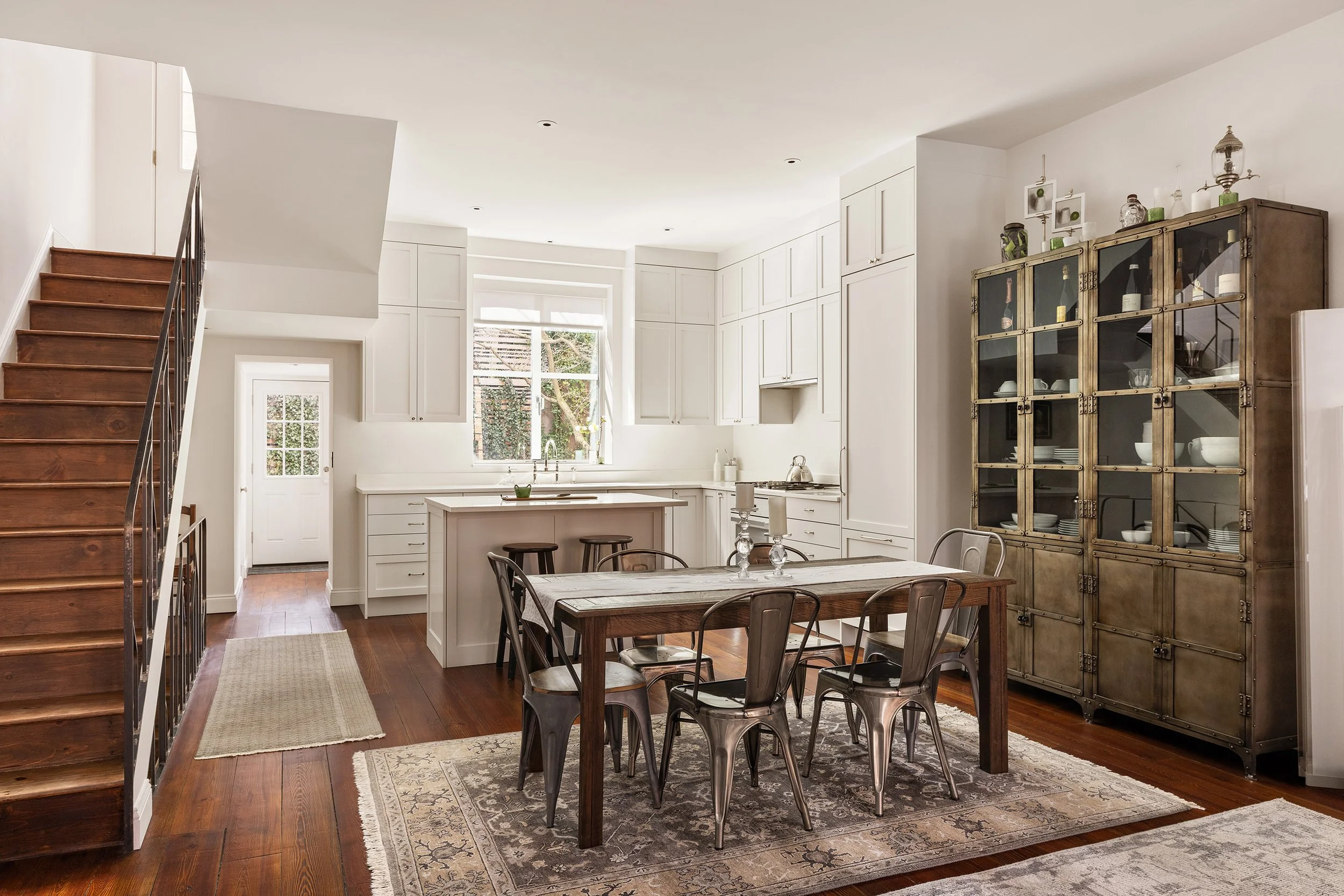 A cozy eat-in kitchen with white cabinets, a wooden dining table with six metal chairs, hardwood floors, a bedroom nearby, a window with a view of trees, and a tall glass-front cabinet filled with dishes and decorative items.