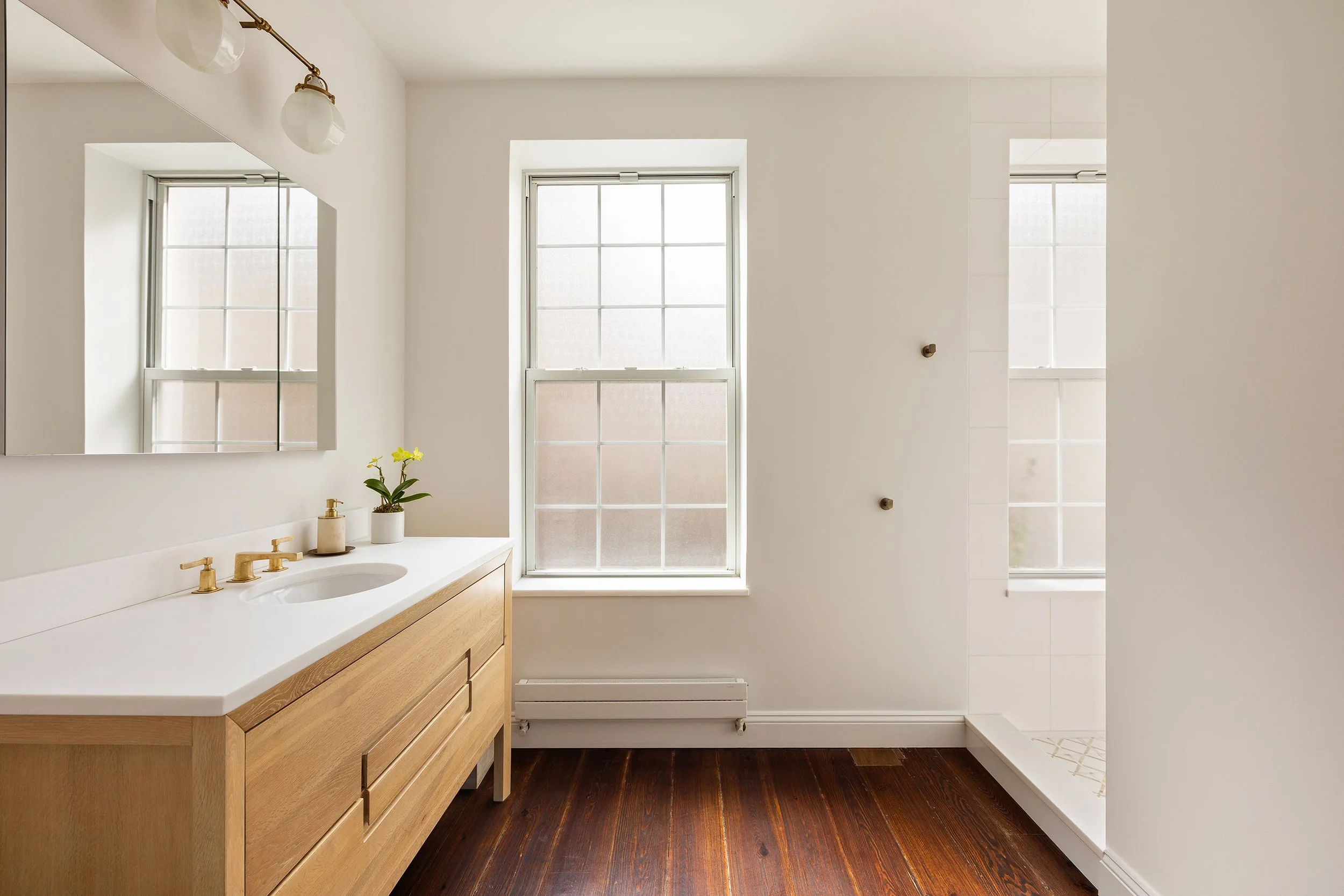 Bathroom with wooden vanity and white countertop, mirror, two windows, and a glass shower enclosure.