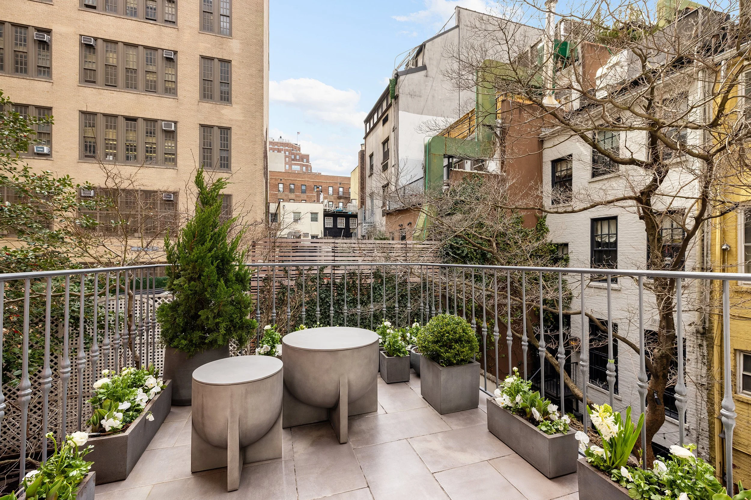 City balcony with metal railing, round concrete tables, potted evergreen trees, and planters with flowers, overlooking neighboring buildings and leafless trees.