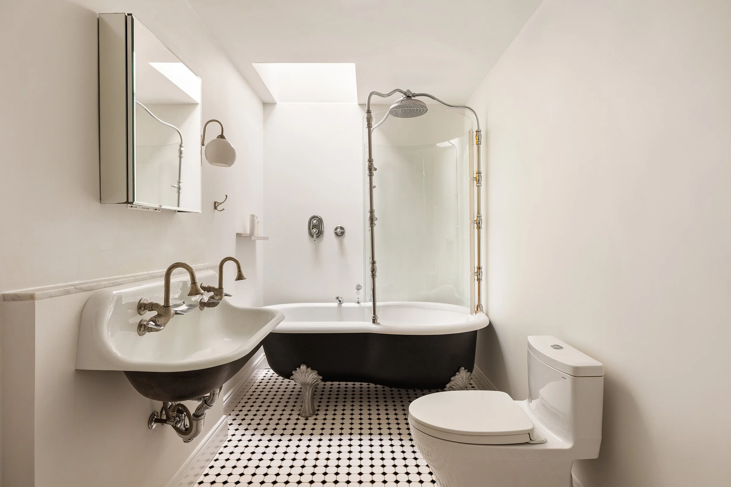 A vintage-style bathroom with a black clawfoot bathtub, wall-mounted sink with brass faucets, modern toilet, a mirror cabinet, a wall-mounted light, a small shelf, and a skylight providing natural light, with black and white hexagonal floor tiles.