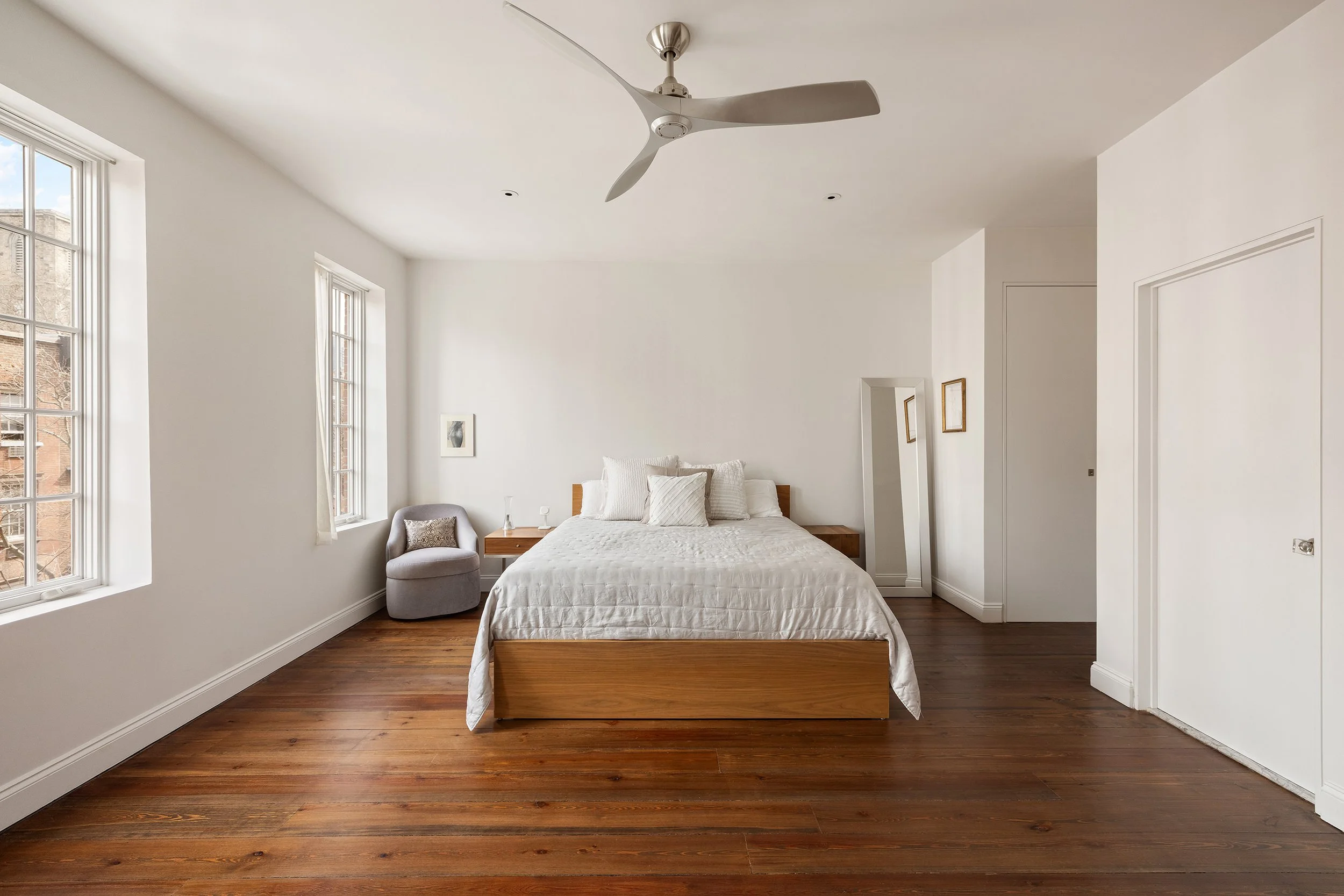 A minimalist bedroom with white walls, a wooden bedframe, white bedding, and a ceiling fan. There are two windows, a gray chair, and a full-length mirror.