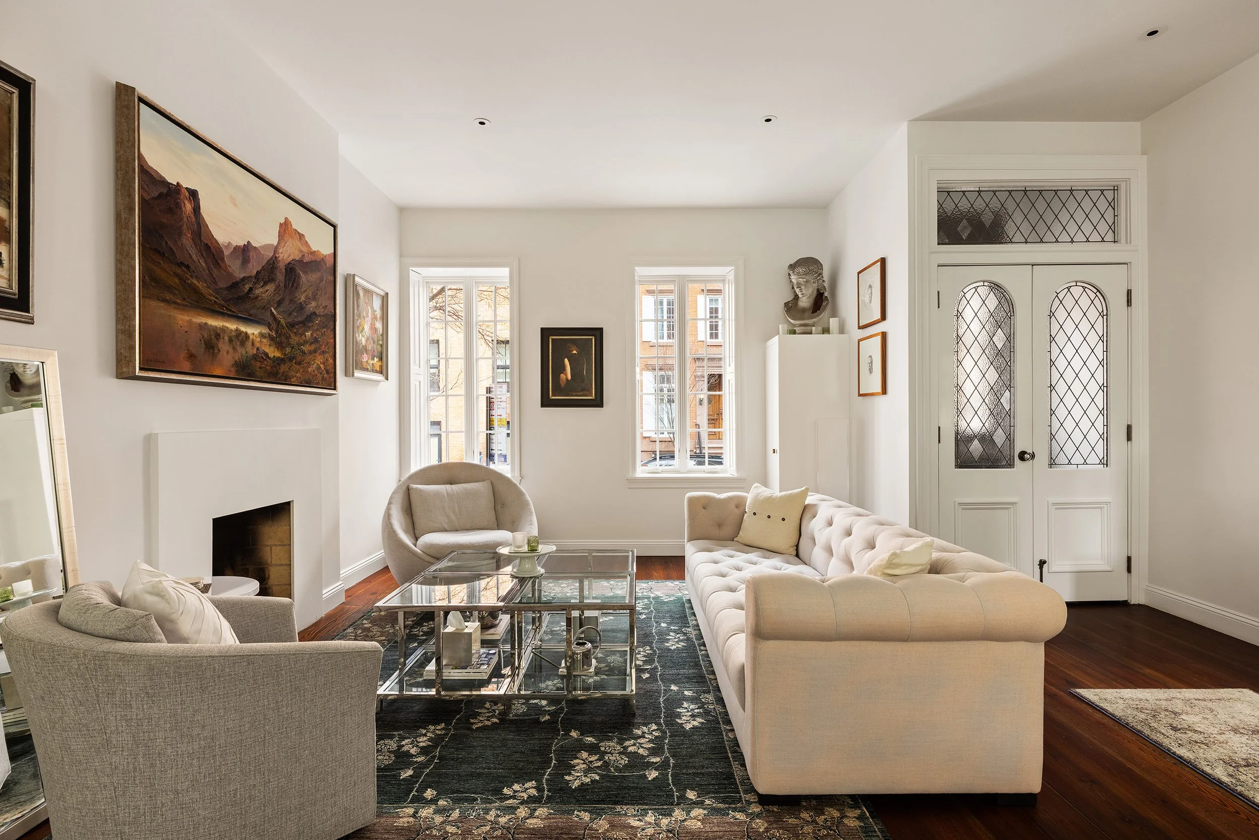 Living room with cream-colored tufted sofa, two armchairs, glass coffee table, artworks on white walls, hardwood floors, and a white door with stained glass.