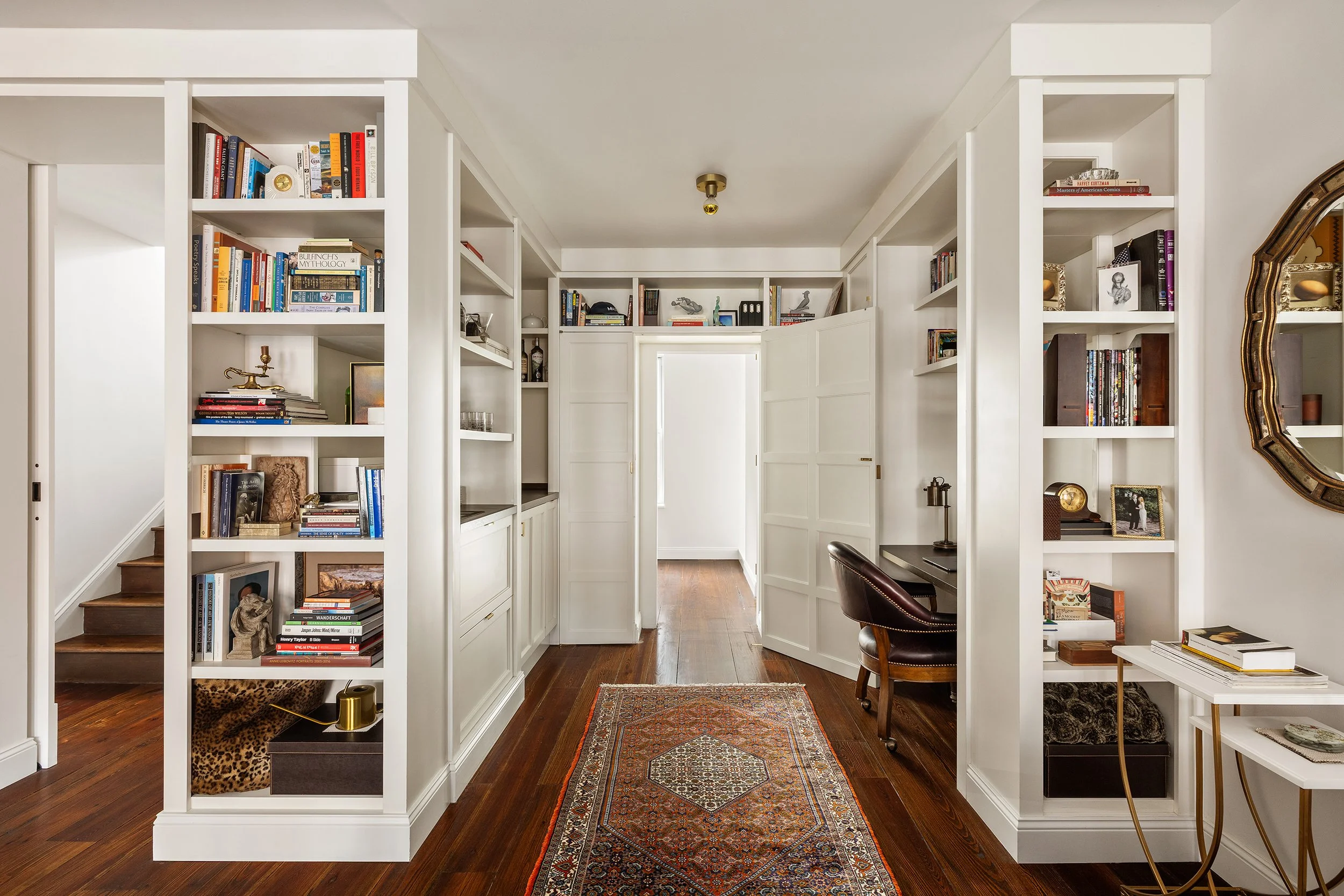 A cozy home office with white built-in bookshelves filled with books, decorations, and photo frames. A dark leather chair sits in front of a small desk, and a colorful patterned rug lies on the polished wooden floor.