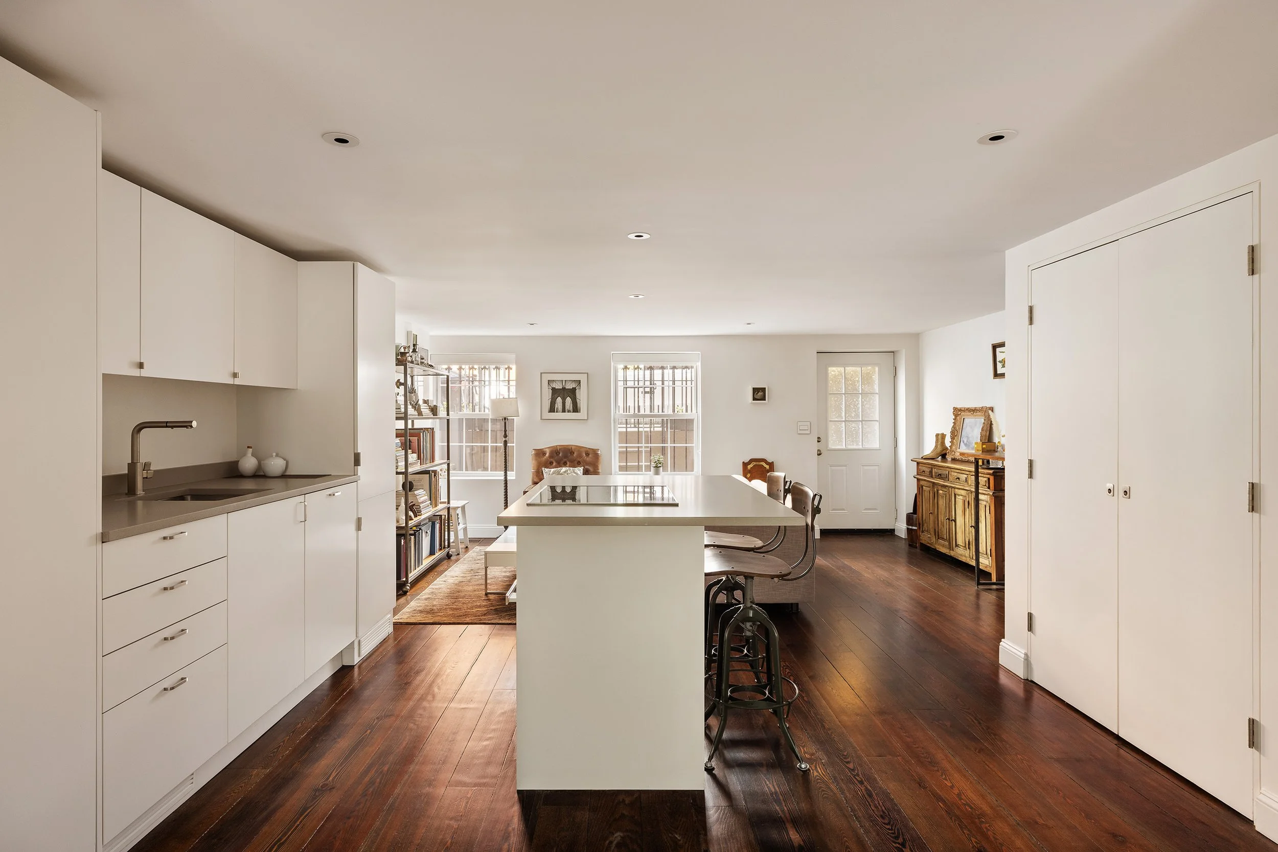 The image shows a spacious, modern kitchen and living area with white cabinetry, a kitchen island, and wooden furniture, illuminated by natural light from several windows.
