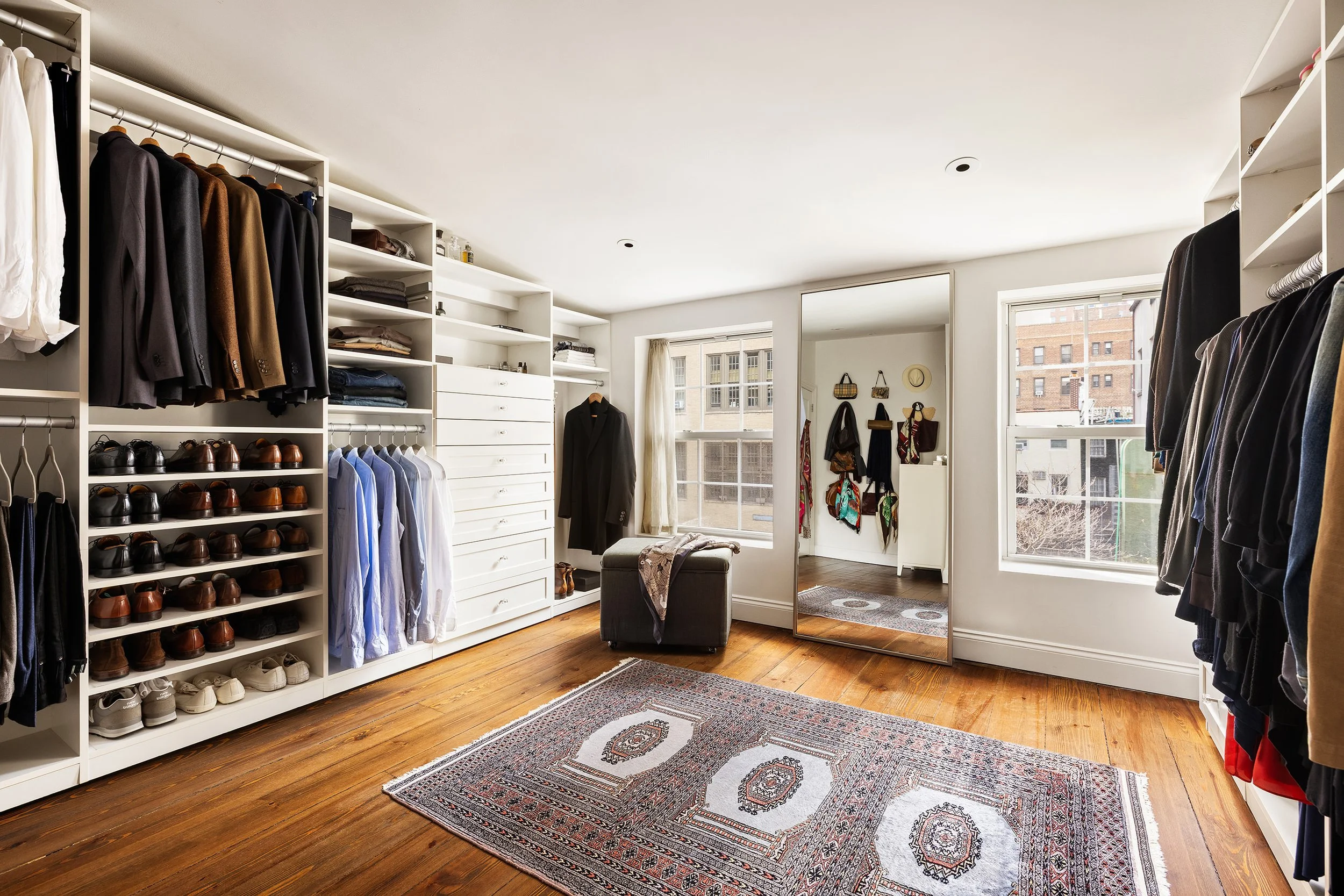 A walk-in closet with white shelves and hanging clothes, shoes on shelves, a full-length mirror, a window showing city buildings, and a wooden floor with an area rug.