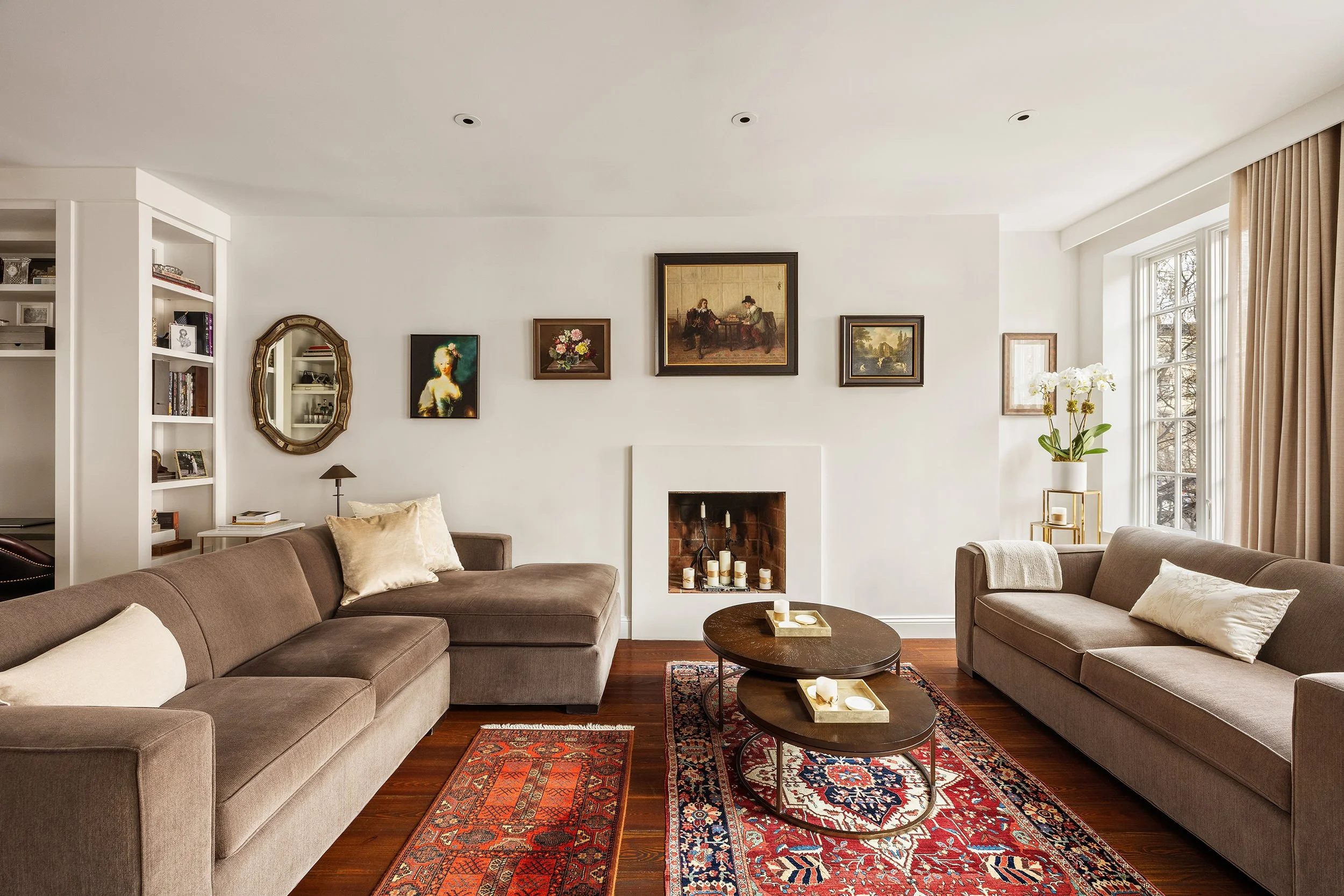 Living room with two beige sofas, a fireplace, artwork on the white wall, a window with curtains, a side table with a plant, and two dark wooden coffee tables on a patterned rug.