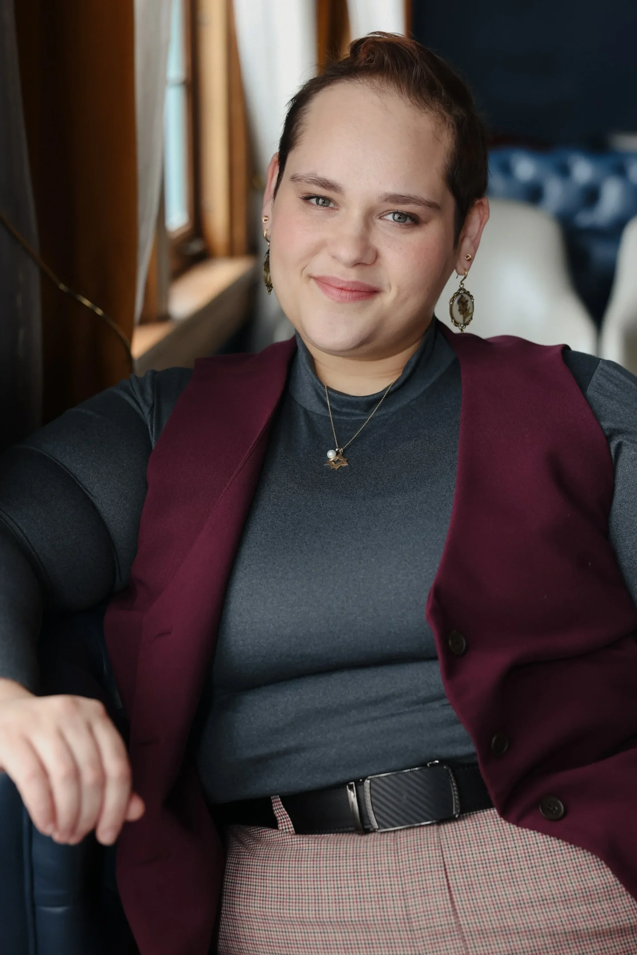 A person with short dark hair sitting in a chair, wearing a dark gray turtleneck, a maroon vest, and patterned pants, smiling at the camera.