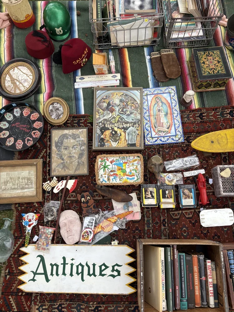 A table covered with various antiques and collectibles, including framed pictures, a painted box, a small bookcase, decorative figurines, and vintage items, with a sign reading 'Antiques'.