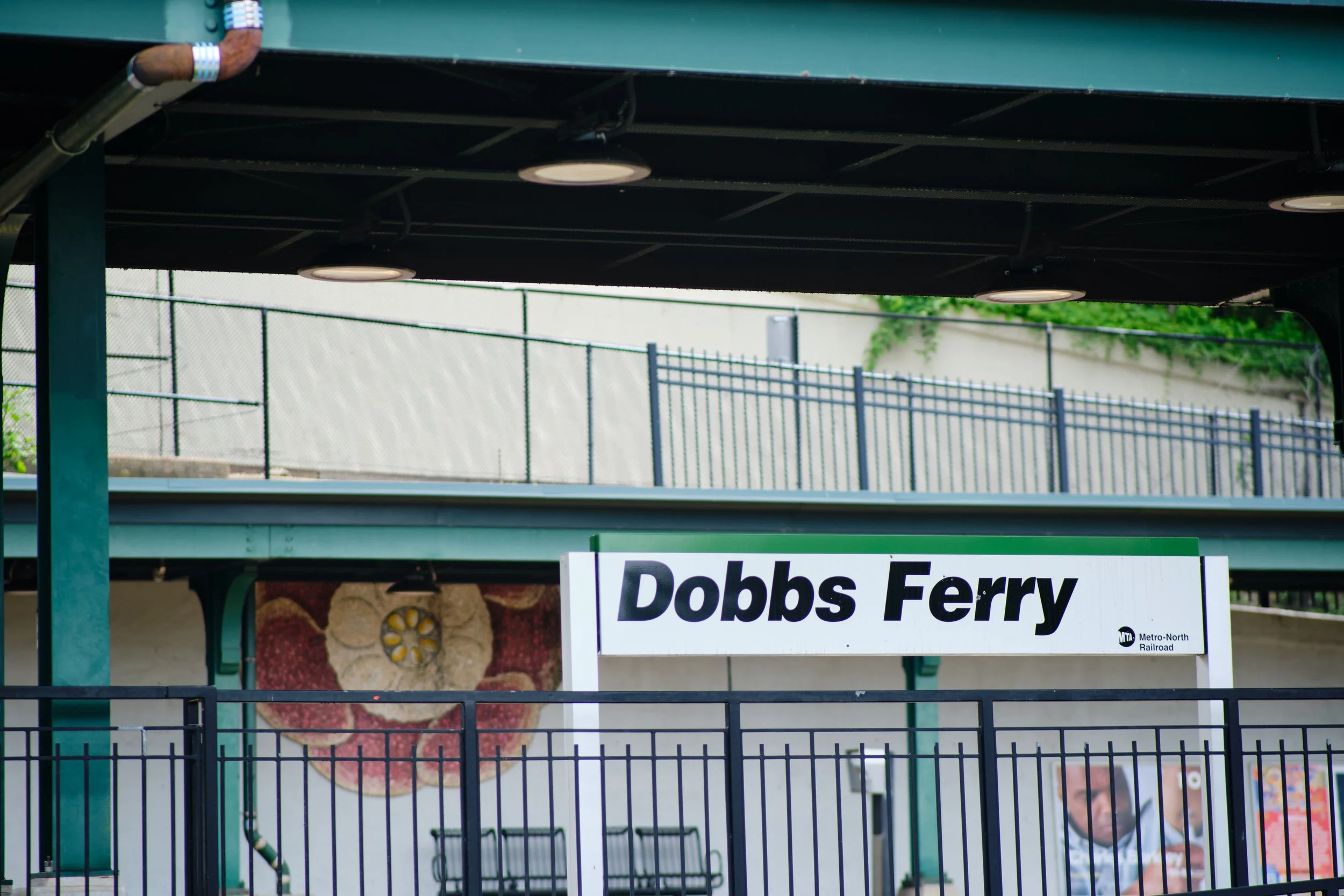 Sign for Dobbs Ferry station of Metro-North Railroad partially visible with a green metal structure above and a black fence in the foreground.
