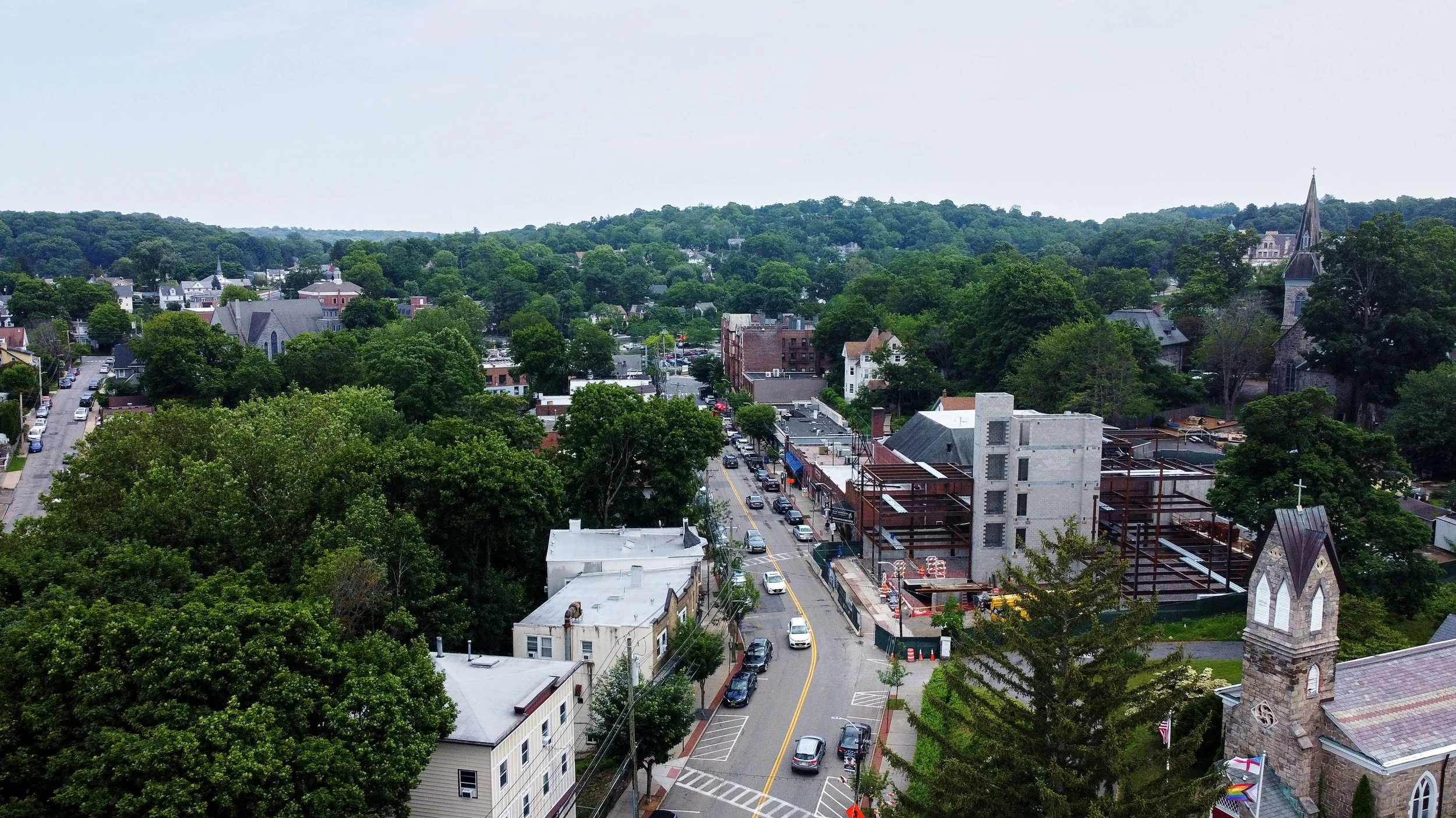 Aerial view of a small town with trees, streets, and buildings, including a church with a tall spire and a partially constructed building.